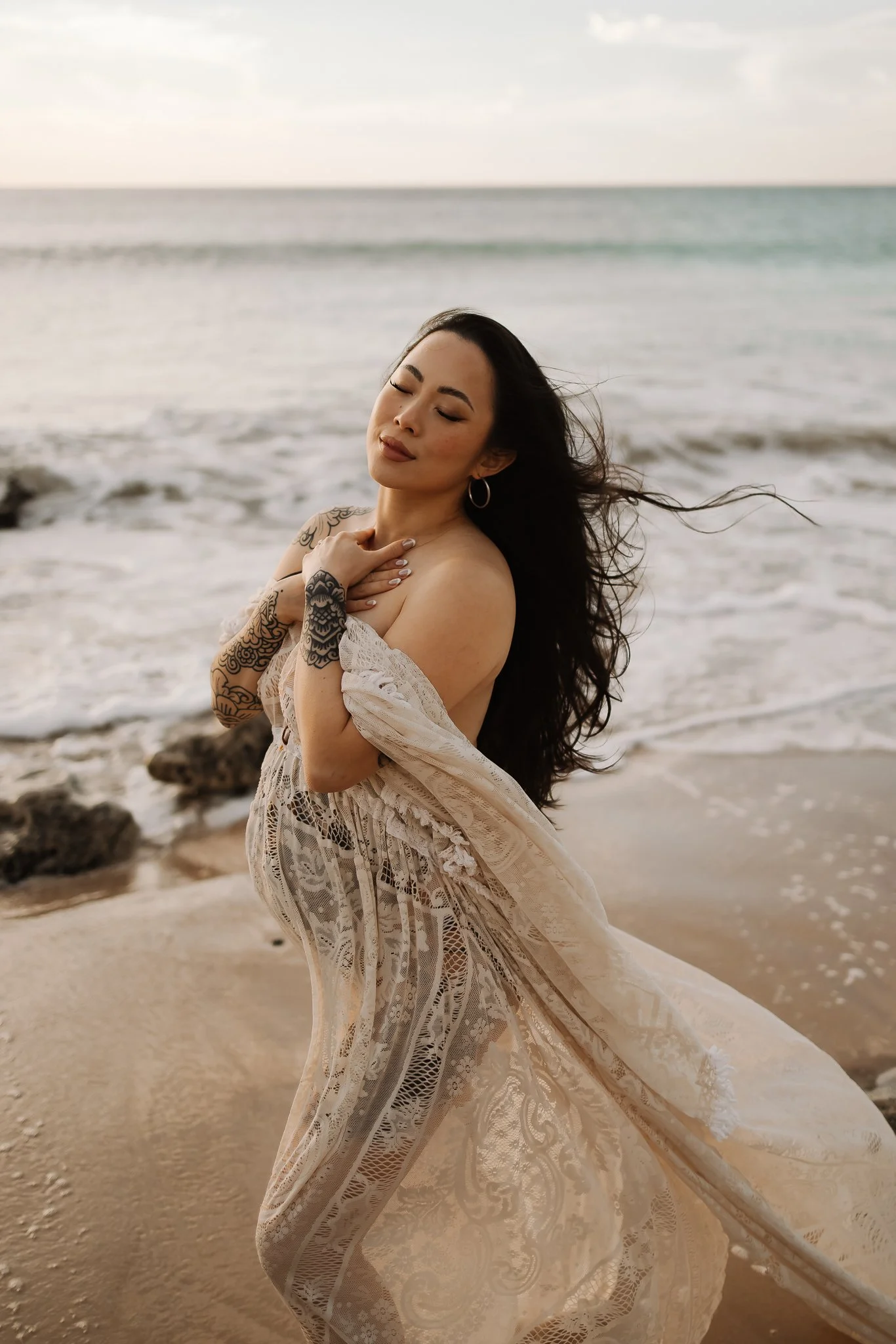 A pregnant Adelaide woman with long dark hair and tattoos on her arms, wearing a light lace dress, standing on a sandy beach near rocks with the ocean in the background, eyes closed and arms crossed over her chest.