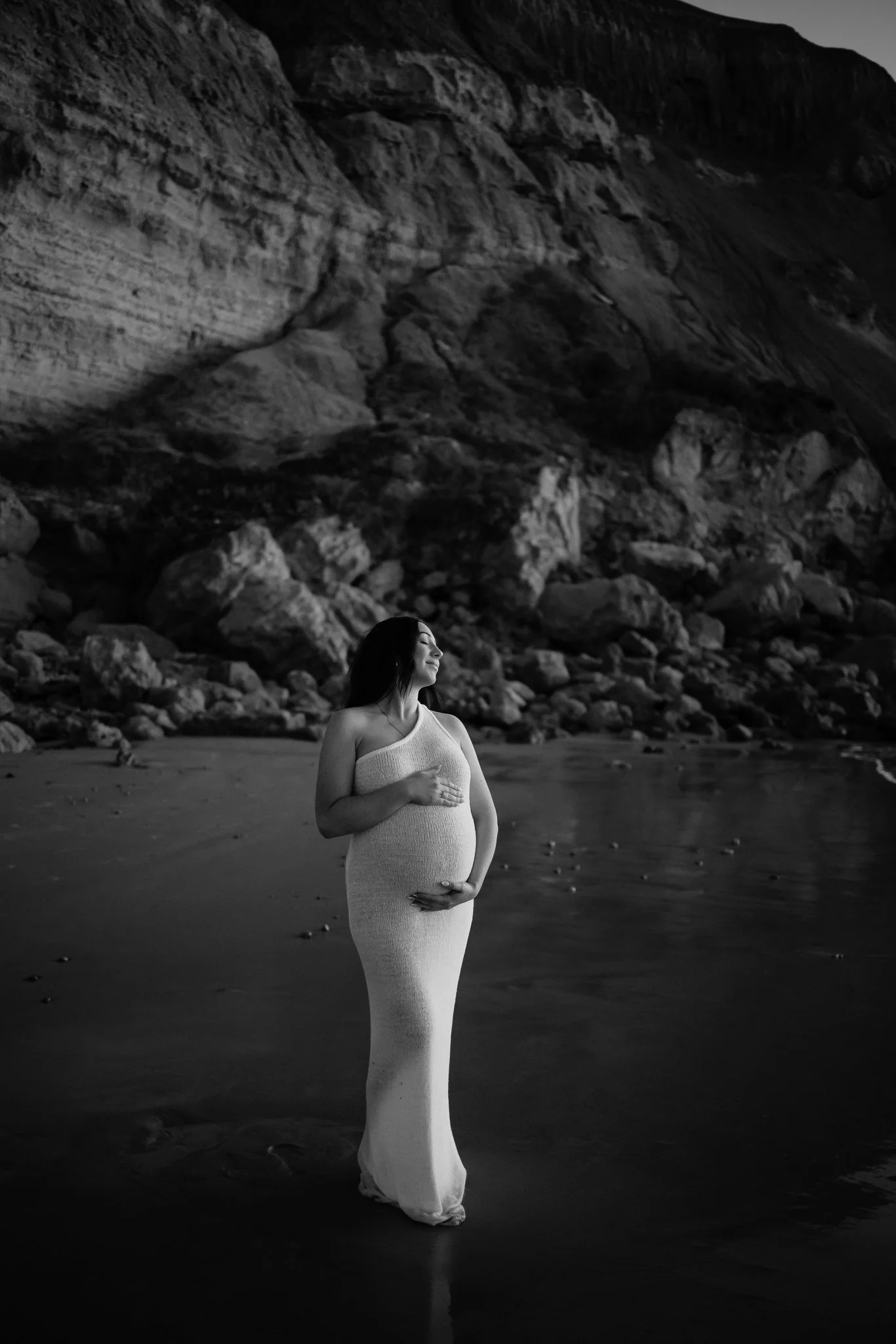 A pregnant woman in a fitted long white knit dress stands on an Adelaide beach, with her face turned upward and her hand on her stomach, near rocky cliffs of Maslin Beach.