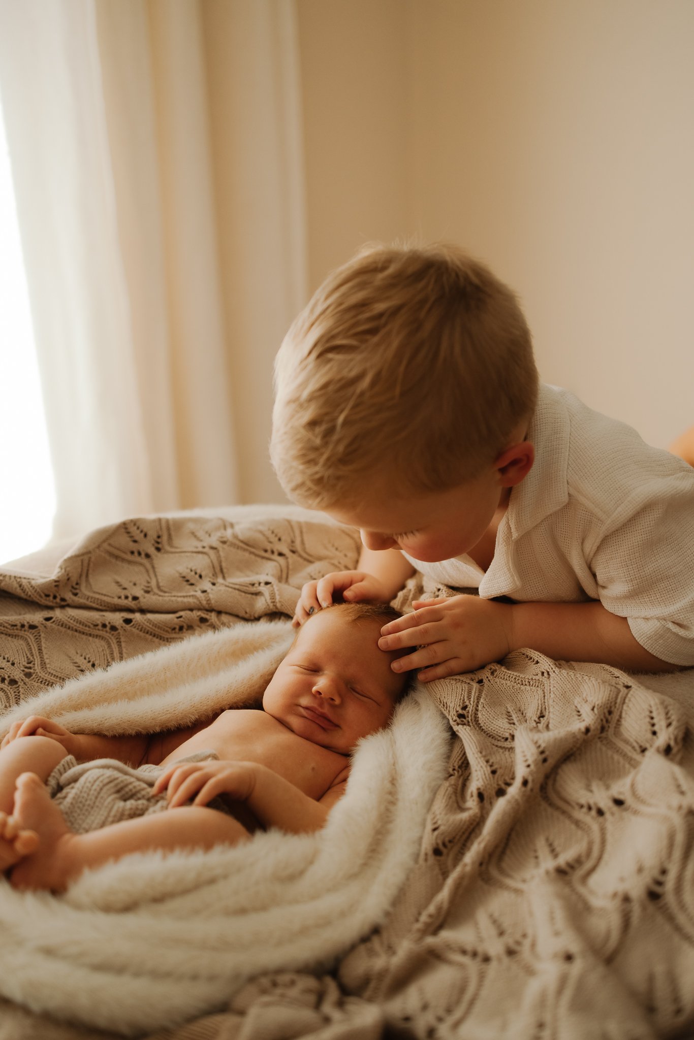A young Adelaide boy leaning over a newborn baby wrapped in a blanket, gently touching the baby's head, in a cozy, softly lit bedroom. Newborn photos at home.