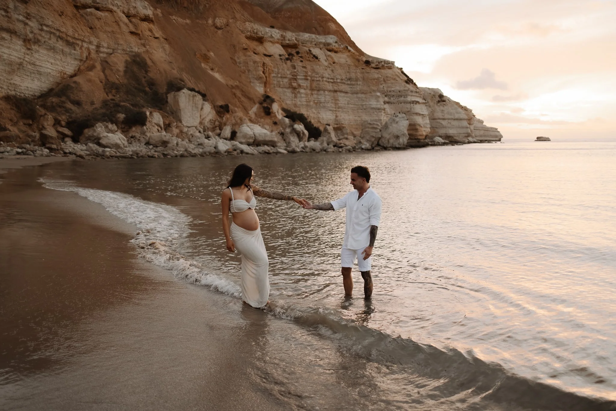 A pregnant couple is standing in the shallow water at the beach during sunset, holding hands, with Adelaide beach cliffs in the background.
