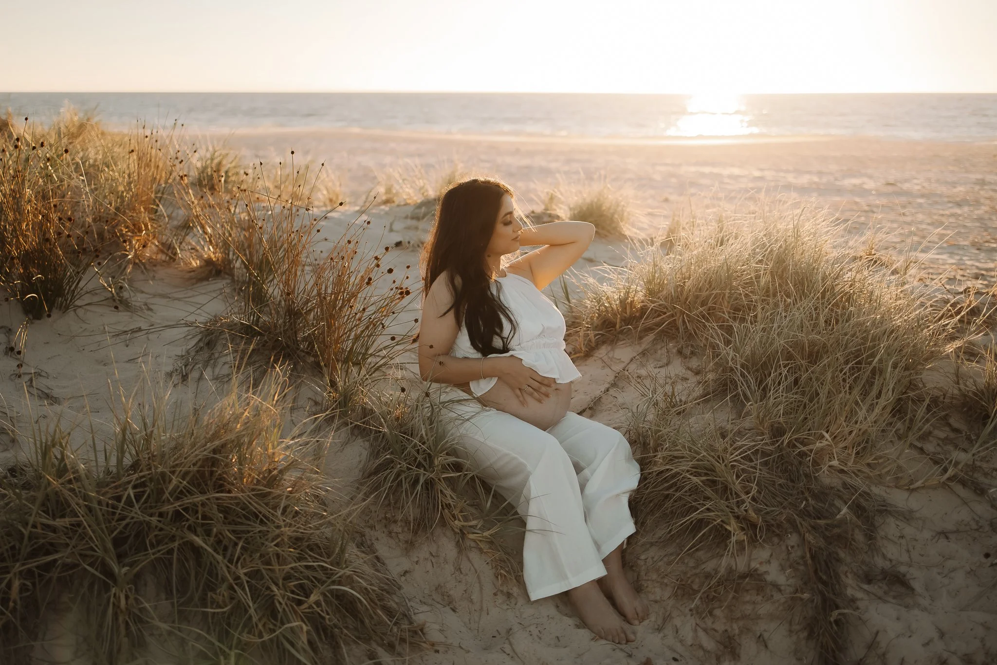 pregnant women sits amongst beach scrub and dunes over looking Port Willunga beach, South of Adelaide at sunset.