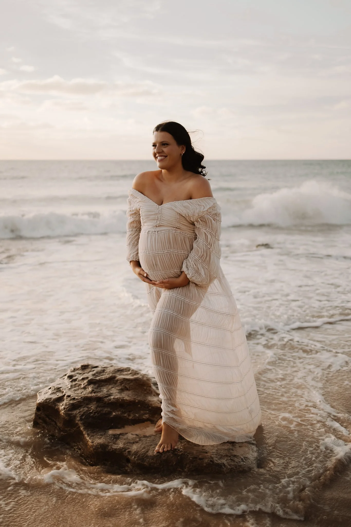 A pregnant woman in a flowing beige dress standing barefoot on a rock an Adelaide beach near the ocean with waves and clouds in the sky, smiling and holding her belly.