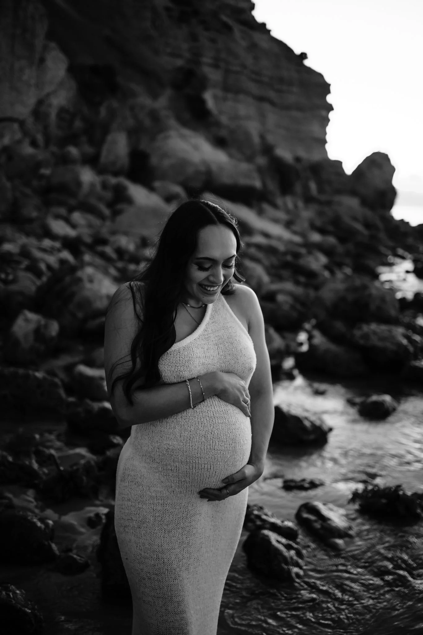 A pregnant woman in a sleeveless, white knit dress stands on a rocky Adelaide beach, smiling and cradling her belly with one hand and resting her other hand on her chest, during sunset or sunrise. Black and white photograph.