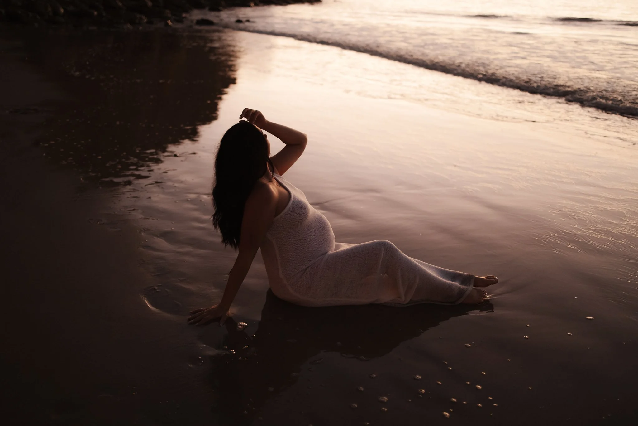 A woman sitting on the wet sand of an Adelaide beach at sunset, with the shoreline and ocean waves visible in the background for her intimate and soulful maternity photoshoot.