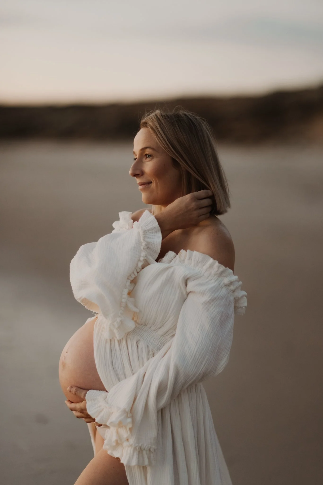 Pregnant woman in a cream-colored dress from Adelaide photographers client closet standing on an Adelaide beach at sunset with her hand gently touching her neck and her other hand supporting her belly.