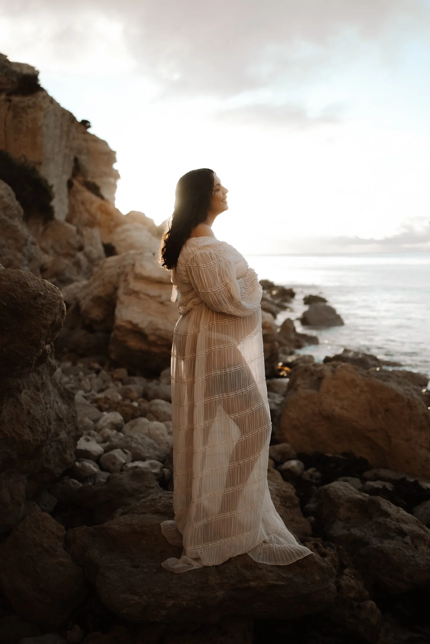 A pregnant woman in a long, sheer, cream-colored dress stands on rocks of an Adelaide beach.. by the ocean at sunset, smiling and looking to the side. The sun shines through the dress making for a dreamy maternity photo.