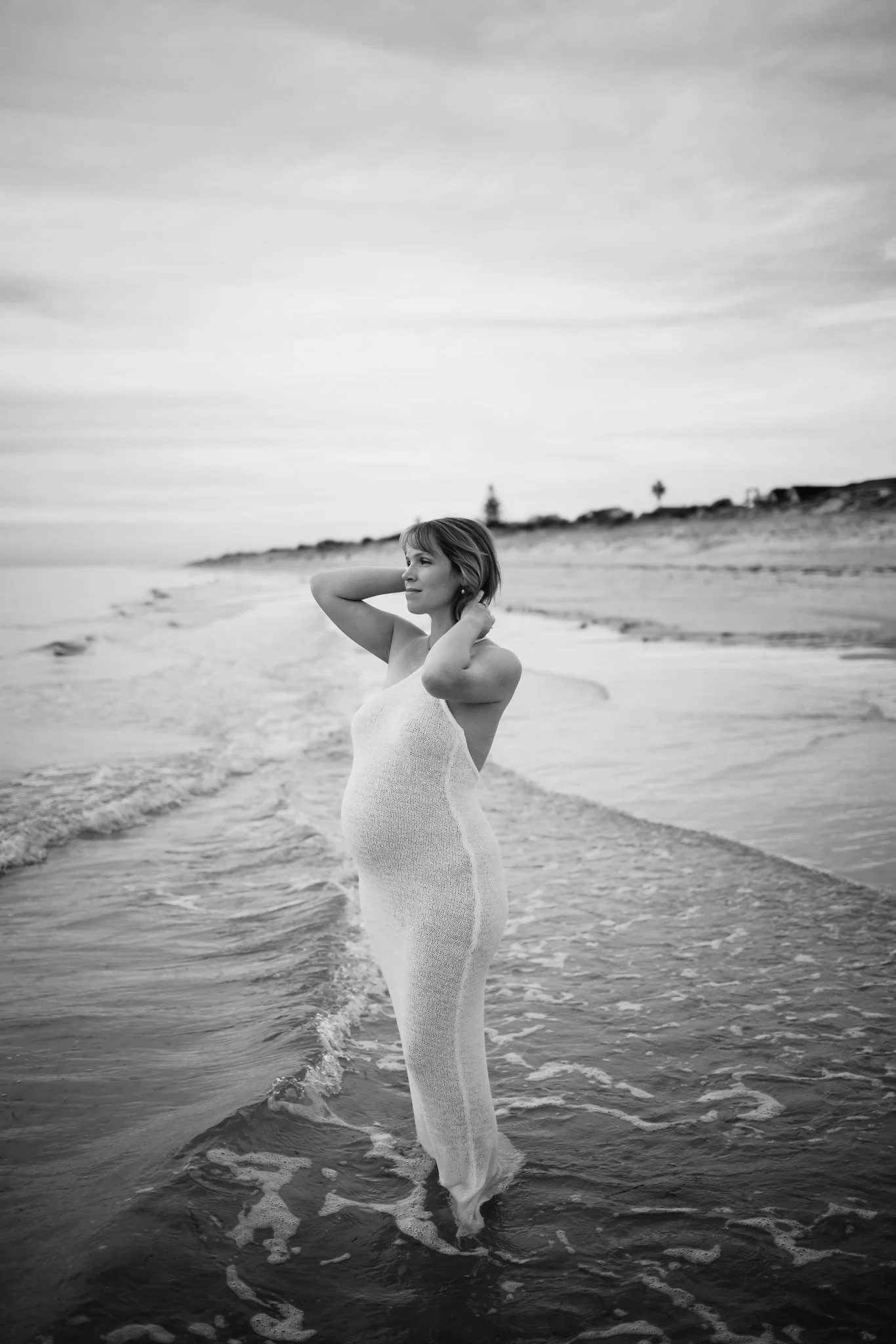 Pregnancy maternity photoshoot at Tennyson Beach, Adelaide. Pregnant woman stands in the water at the beach looking out to the ocean while she adjusts her hair. natural and romantic maternity photos.