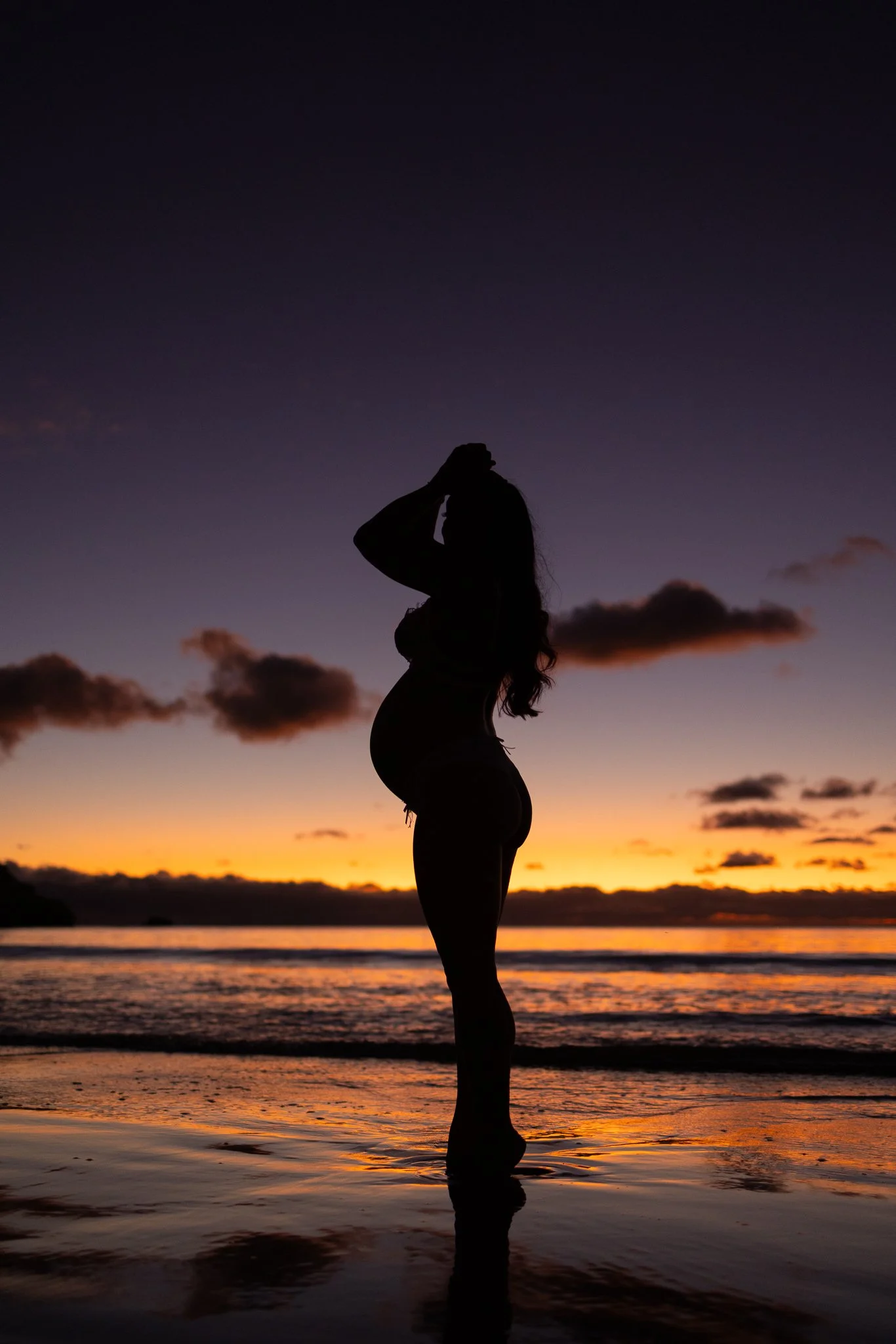 Silhouette of a pregnant woman standing at an Adelaide beach during sunset. The shape of her pregnancy bump is seen as well as the bright colours of the sunset.