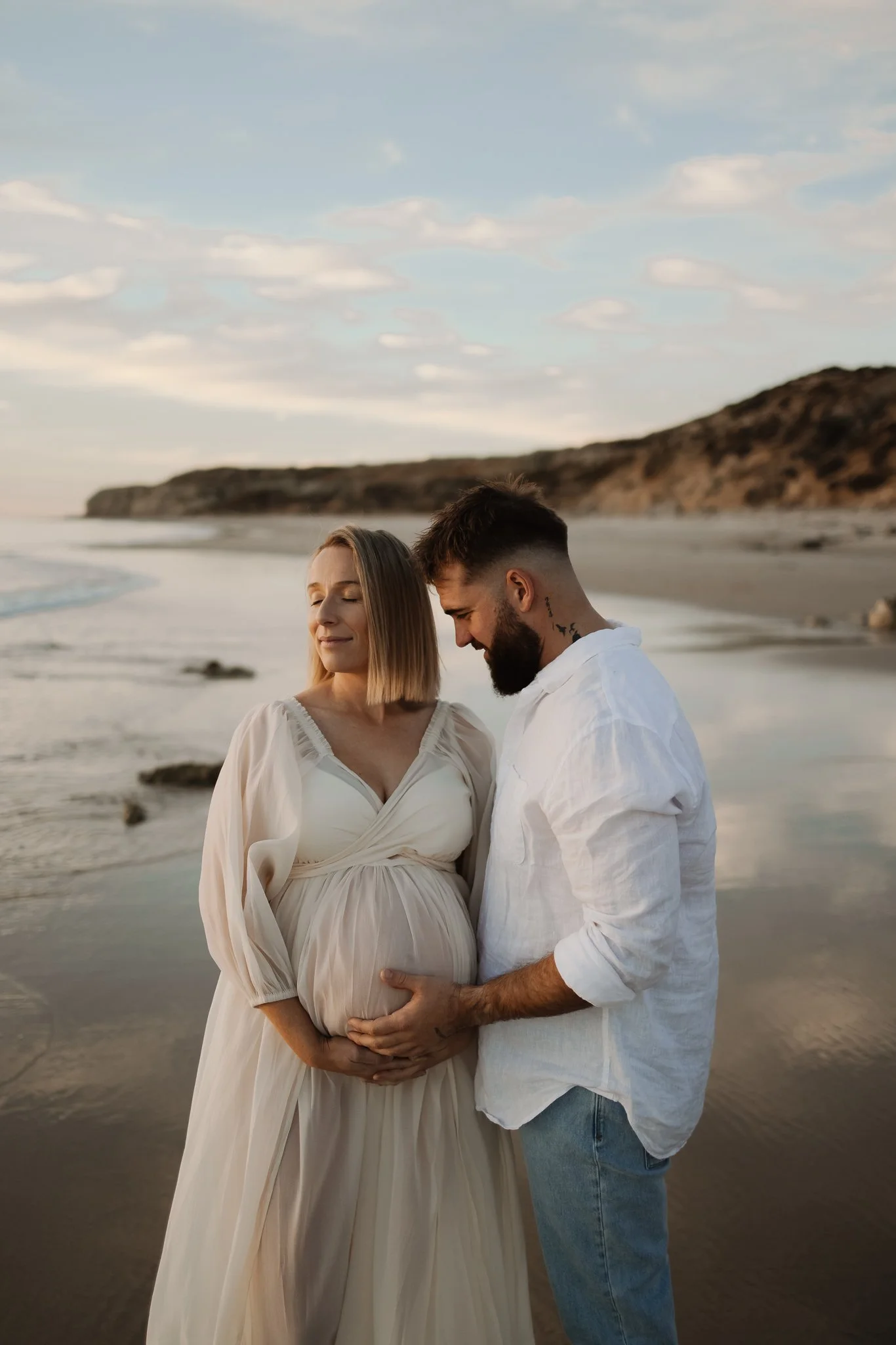 A pregnant woman and a man standing on an Adelaide beach during sunset, with the woman's belly gently held by both of them, as they share an intimate moment for their maternity photoshoot.