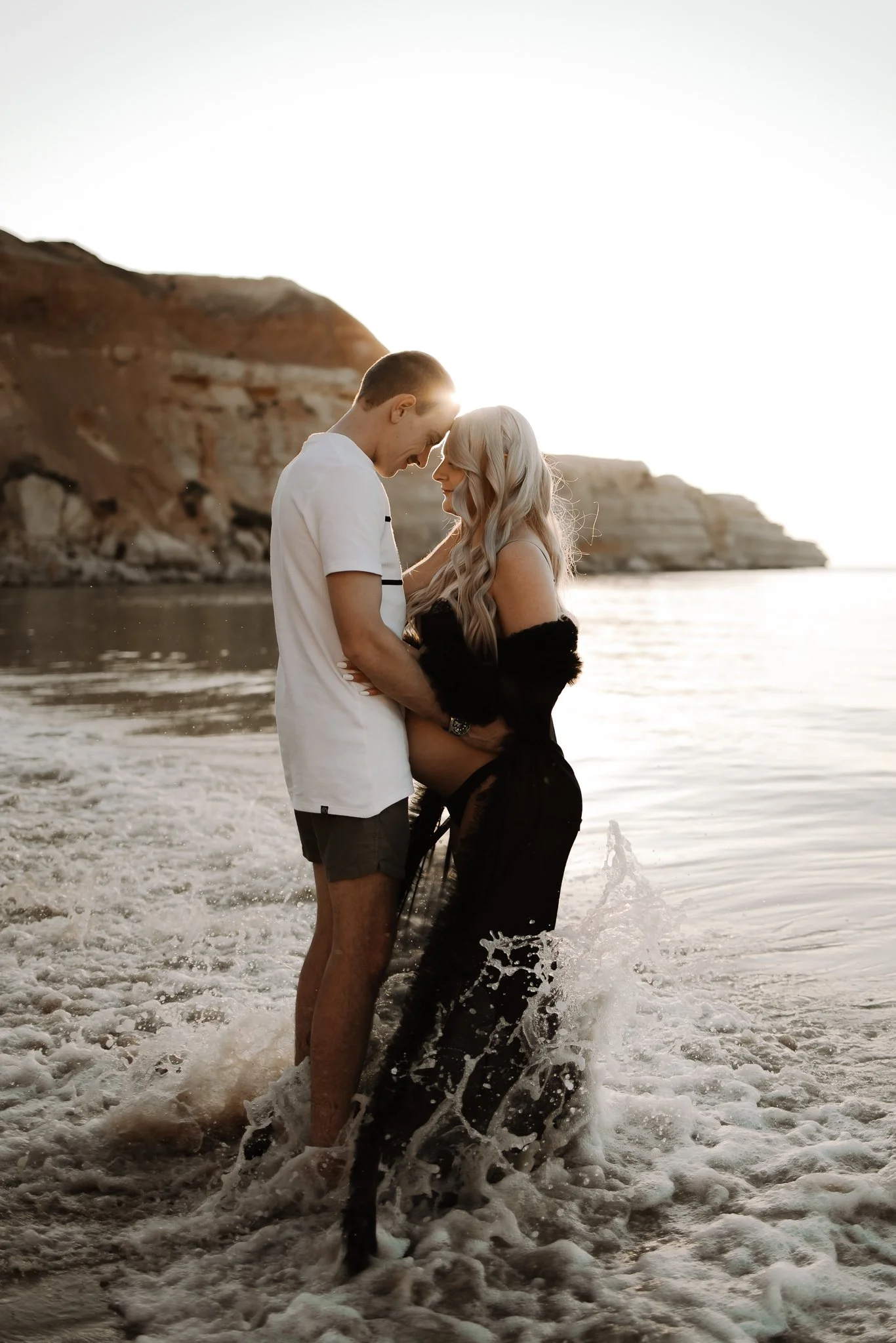 A couple standing in the ocean at sunset, touching foreheads, hands on her pregnant bump with beautiful rocky cliffs in the background at a Adelaide beach during their maternity photos.