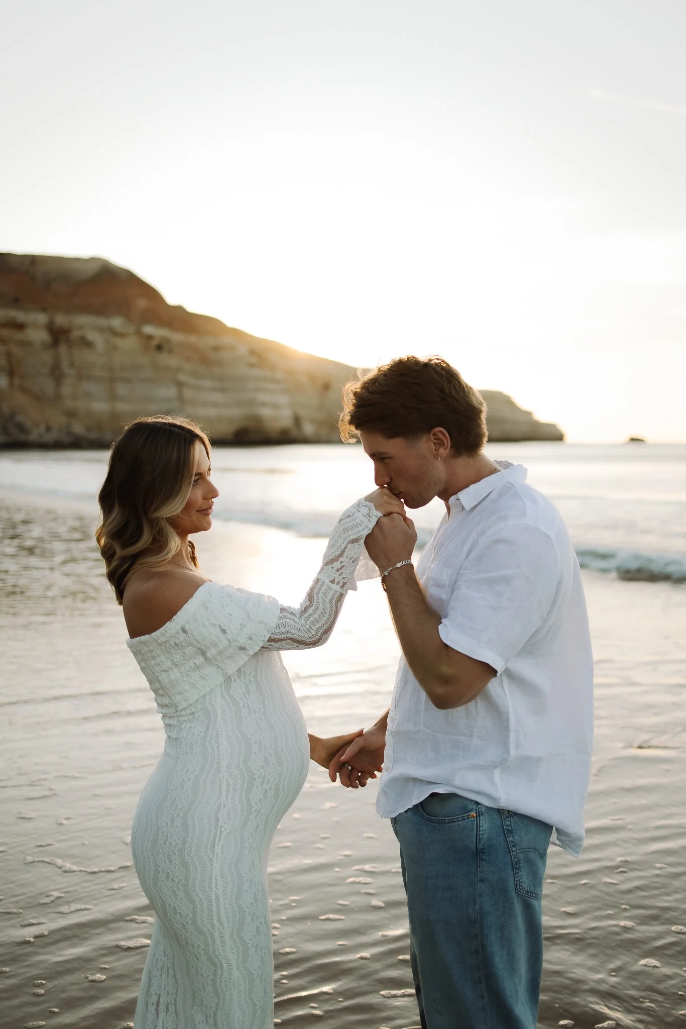 A pregnant woman and a man are on Maslin beach during sunset, holding hands; the woman is kissing the man's hand.