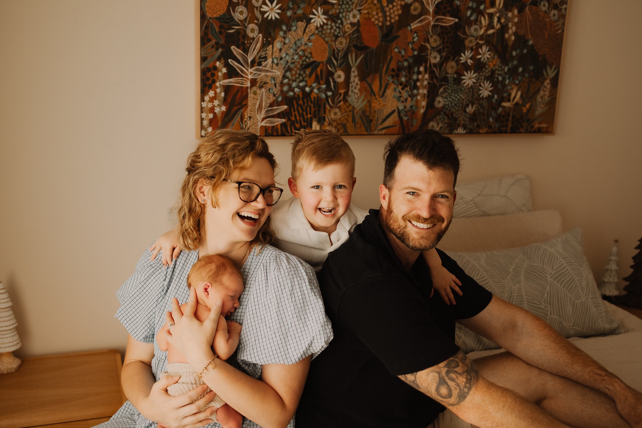 A happy Adelaide family of four sitting on a bed, smiling at the camera. The mother holds a newborn baby, and her hand rests on the toddler sitting on her shoulder. The father is sitting beside them, all in a cozy bedroom with wall art and pillows.