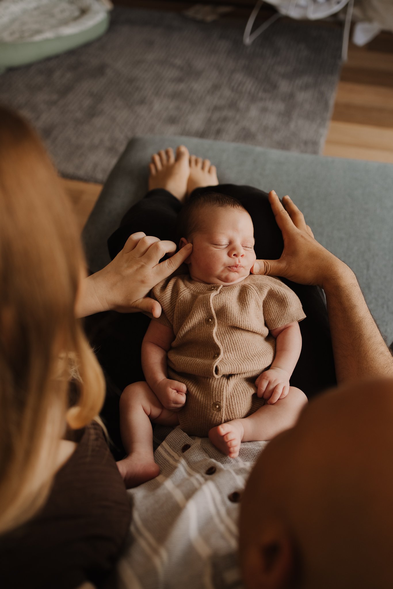 An Adelaide Parent sits on their couch in their home holding their newborn baby on their legs and gently touching his face and admiring him as he lays there all sweet wearing a brown knit romper. 
