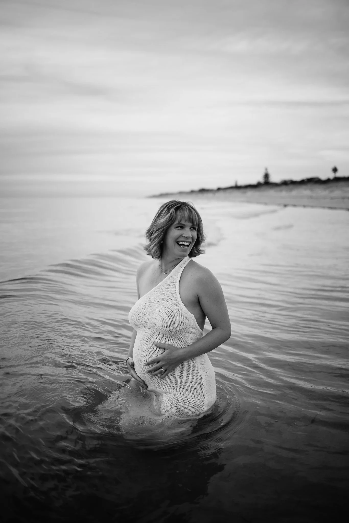 Black and White photo of a pregnant woman smiling while sitting in the ocean water, wearing a sleeveless white knit dress, with an Adelaide beach and cloudy sky in the background.