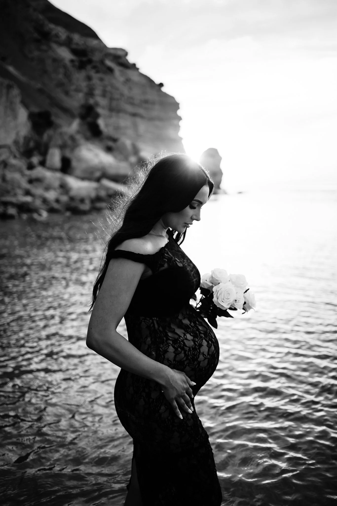 A black and white photo of a pregnant woman in a black lace dress holding a bouquet of white roses standing in the water at an Adelaide beach at sunset, with rocky cliffs in the background.