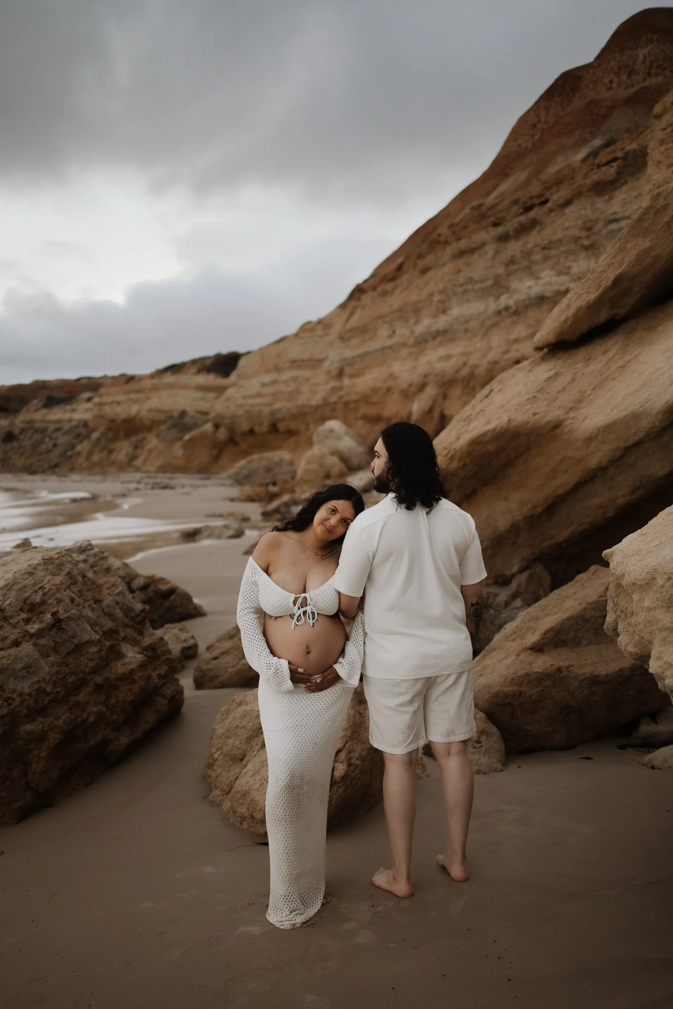Pregnant woman in white crochet dress hugging her belly with partner on an Adelaide beach with rocks and cloudy sky in the background.