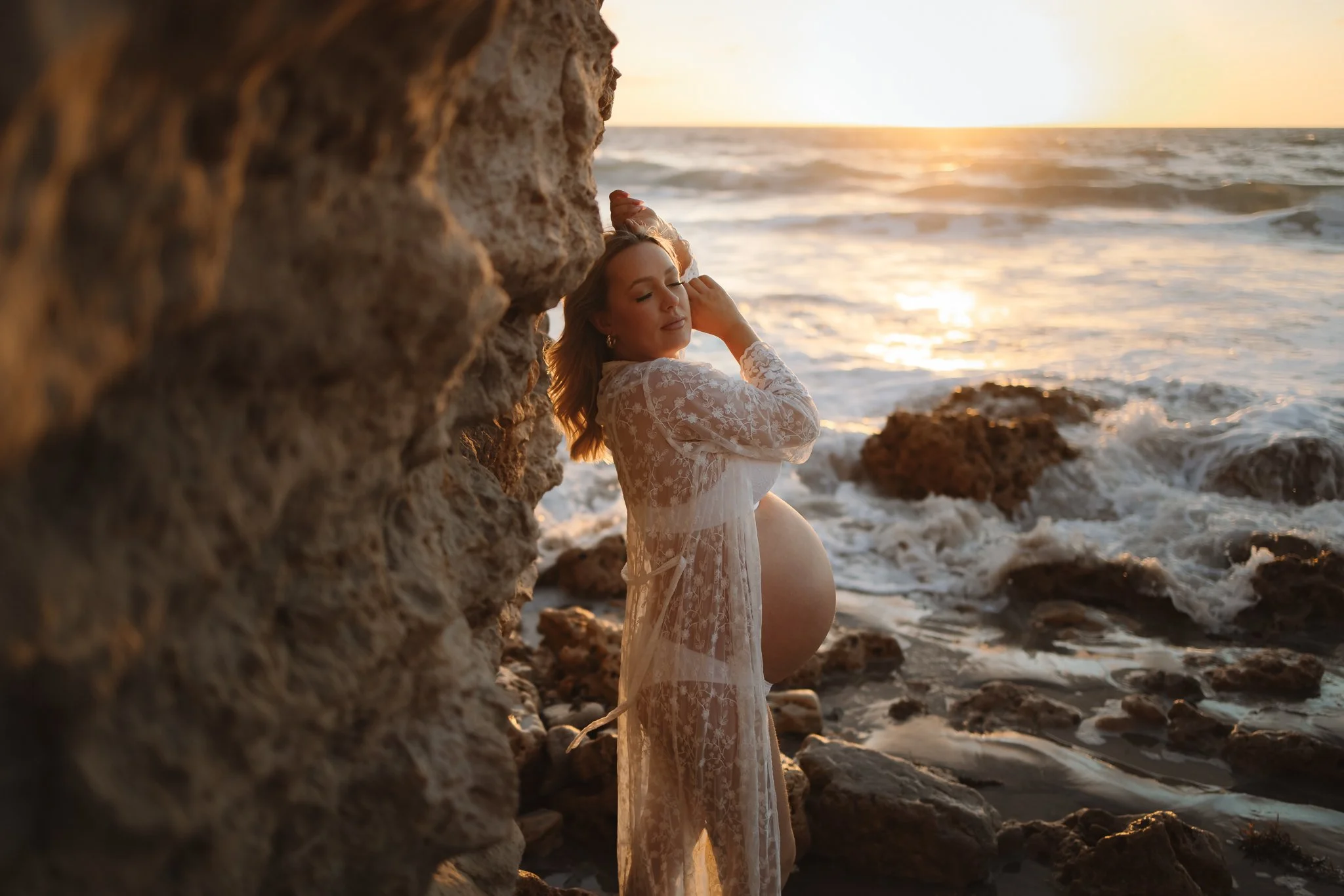 Pregnant woman in a white lace robe standing by rocks on an Adelaide beach at sunset, with her eyes closed and having a dreamy and intimate moment soaking up the last moments of her pregnancy.