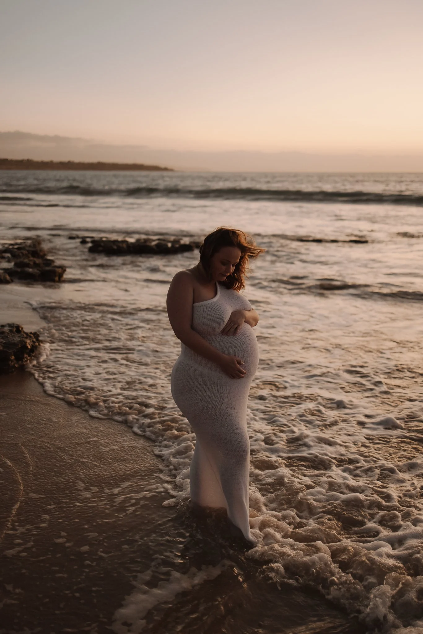 A pregnant woman standing on the shoreline of an Adelaide beach at sunset, wearing a white knit dress and gently hugging her growing belly. The light is soft, just after the sun had set, creating a romantic maternity photo.