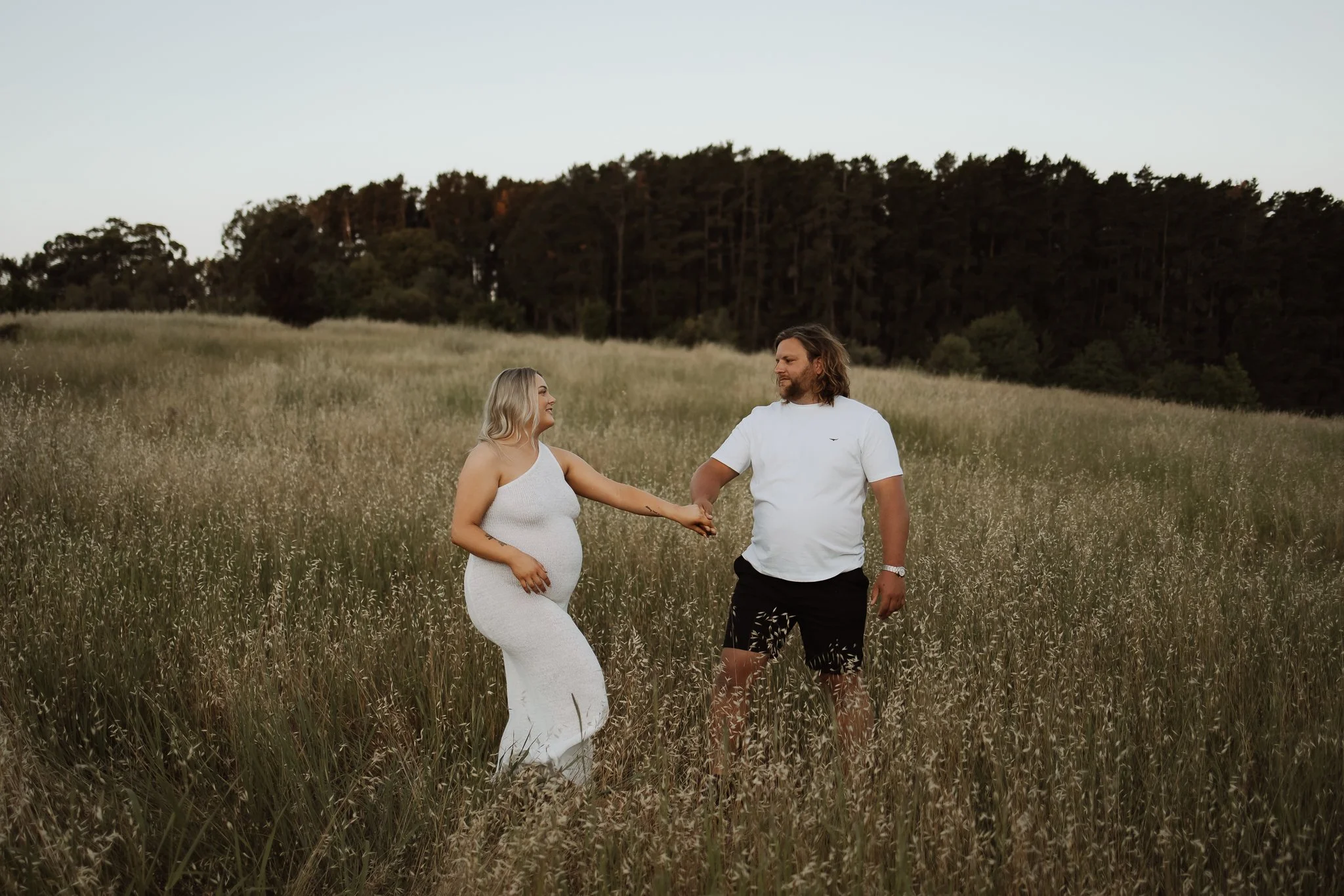 pregnant women walking towards husband in a field with Blackwood forest in the background. romantic and beautiful maternity photography Adelaide.