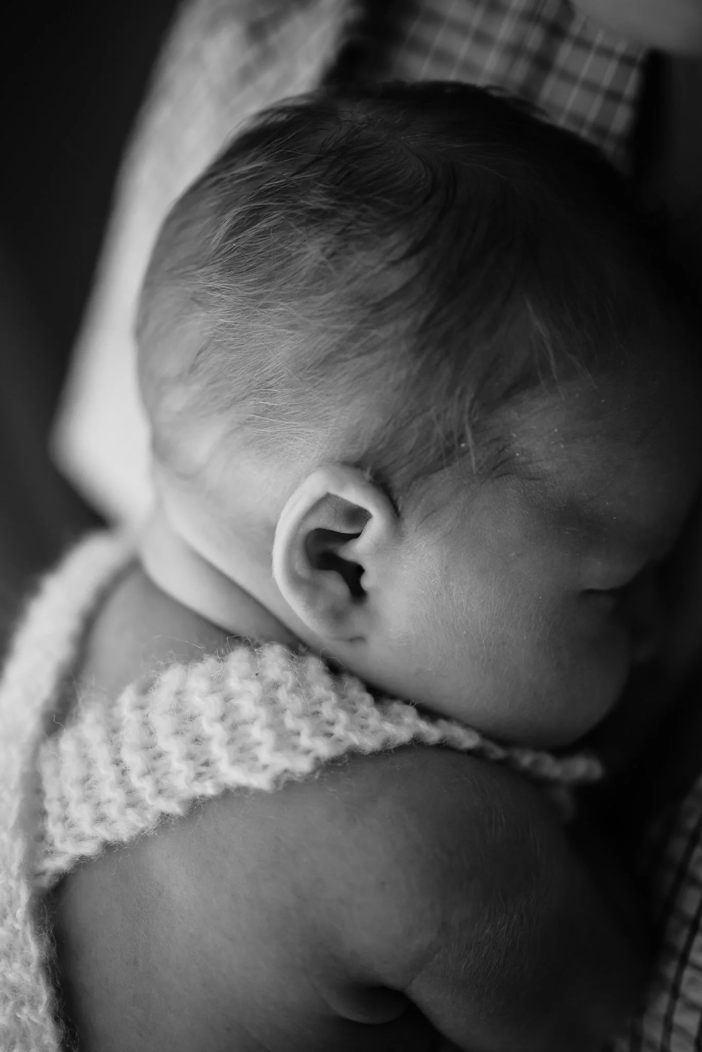 Close-up of a sleeping baby's ear with soft hair, wearing a knitted garment, resting on an adult's shoulder. Beautiful detail photos at your in home newborn photoshoot Adelaide.