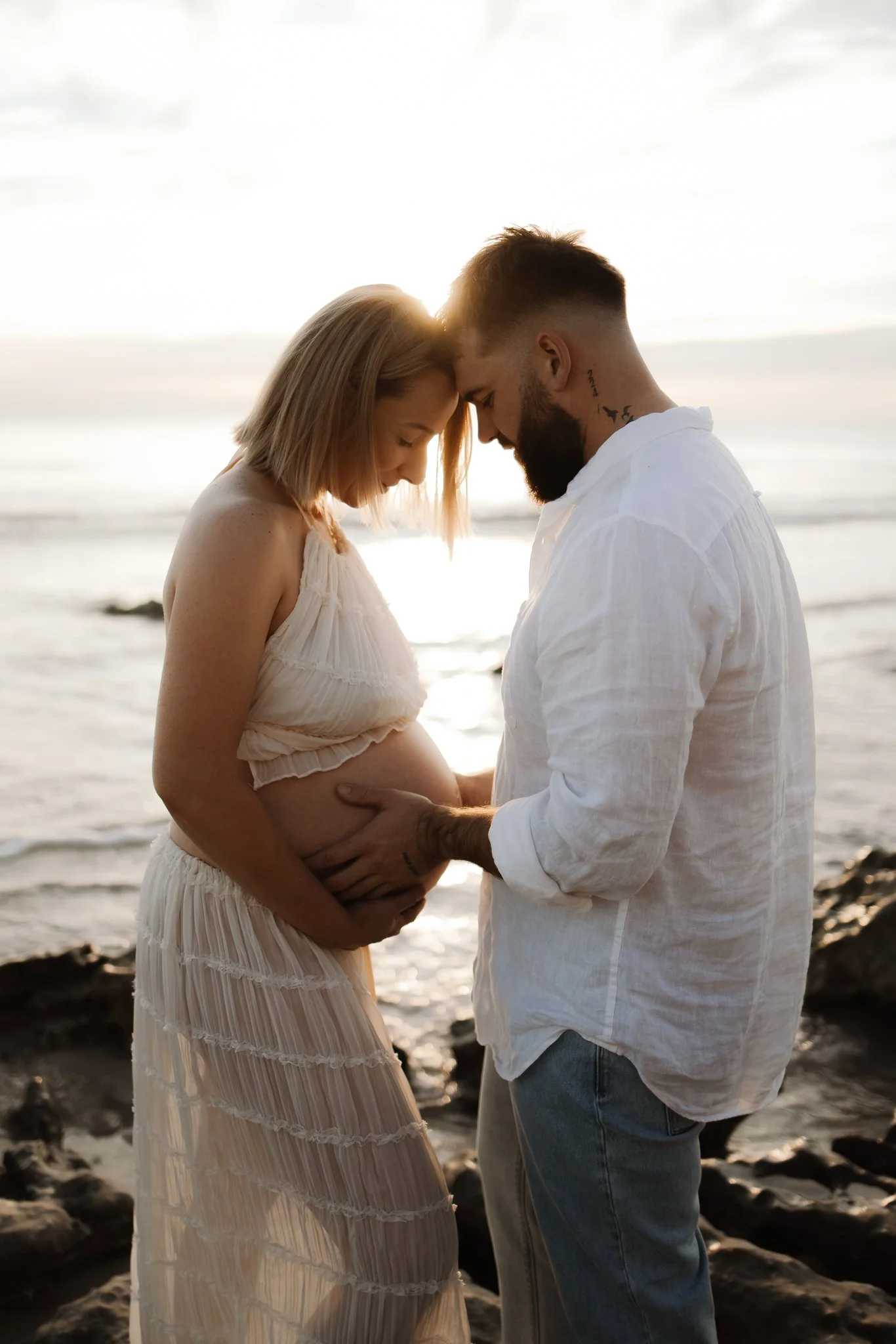 A pregnant woman and a man are standing on an Adelaide beach at sunset, gently touching and looking at her belly with their foreheads pressed together for their Maternity photography.