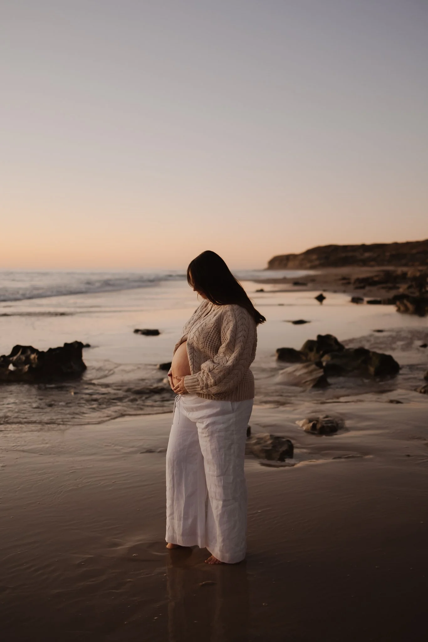 A pregnant woman in white pants and a beige knitted sweater standing barefoot on an Adelaide beach at sunset, cradling her belly for her Maternity photoshoot.