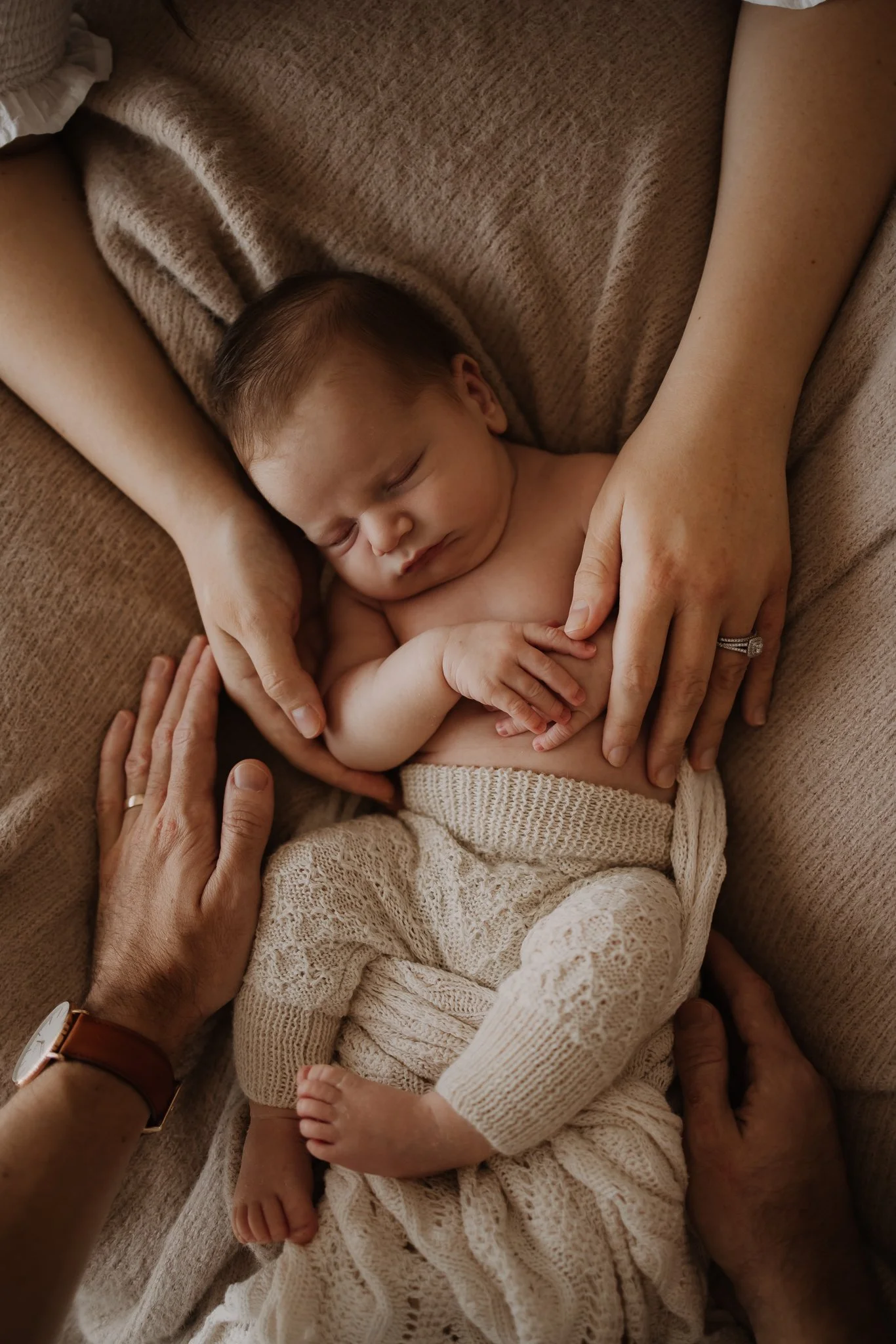 Newborn baby boy in knitted pants sleeping all cozy on blankets with his parents hands surrounding him with love and comfort at their in home newborn session north east of Adelaide, Modbury, Tea tree Plaza.