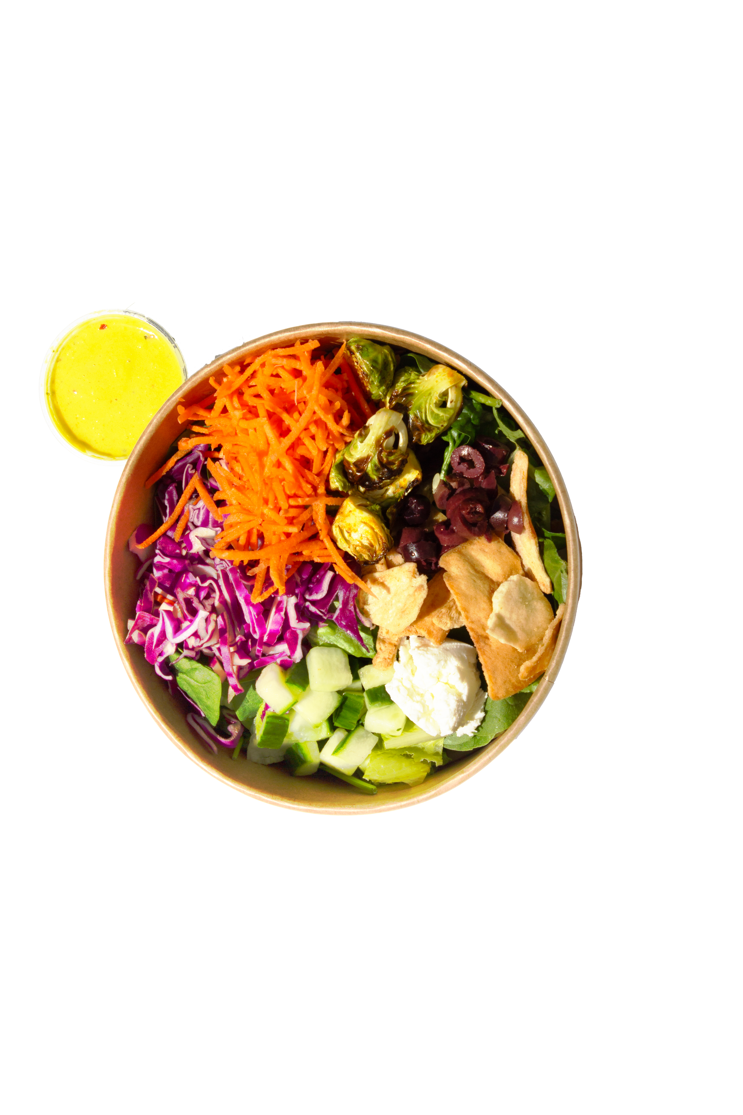 Box of salad with chickpeas, shredded carrots, sliced cucumbers, feta cheese, chopped cucumbers, red cabbage, and a small container of yellow dressing.