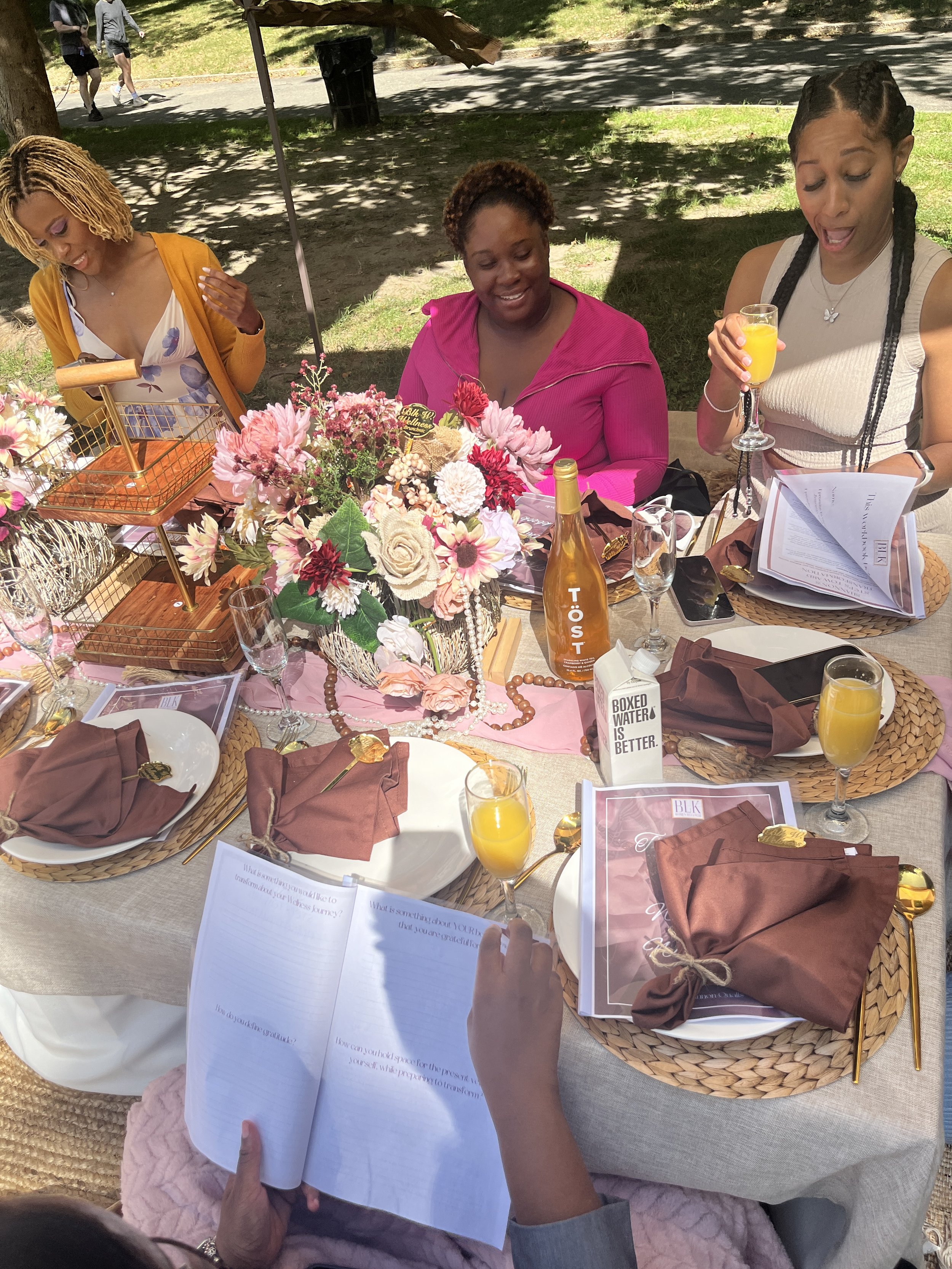 Three women sit at a decorated outdoor table with pink and white flowers, pink table runner, brown napkins, and orange drinks. They are smiling and reading, with one woman holding a glass of orange juice and another with a booklet.