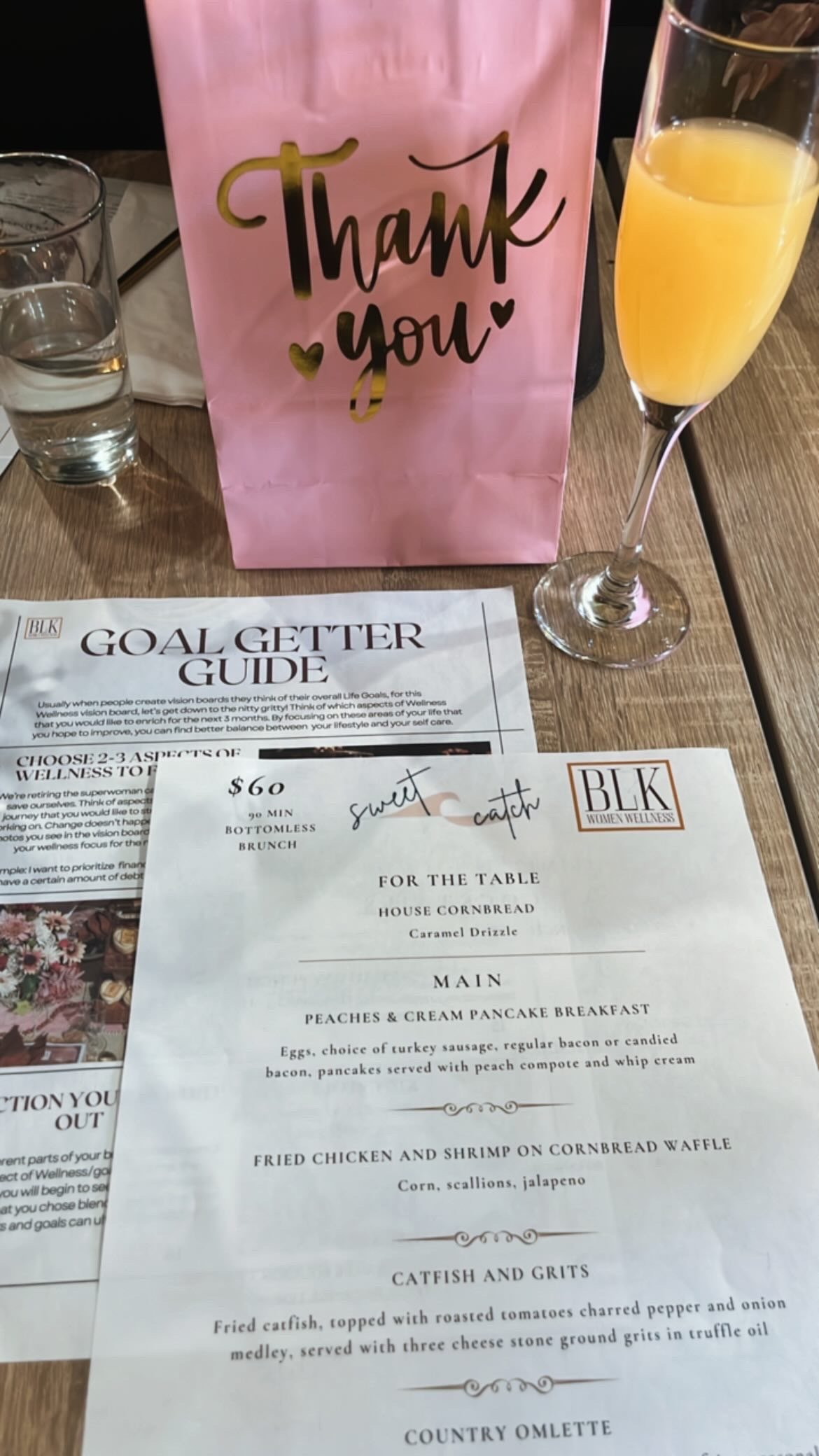 Pink gift bag with gold and black lettering that says "Thank you" surrounded by two small hearts, a glass of orange juice, a glass of water, and a menu on a wooden table at a restaurant or event.