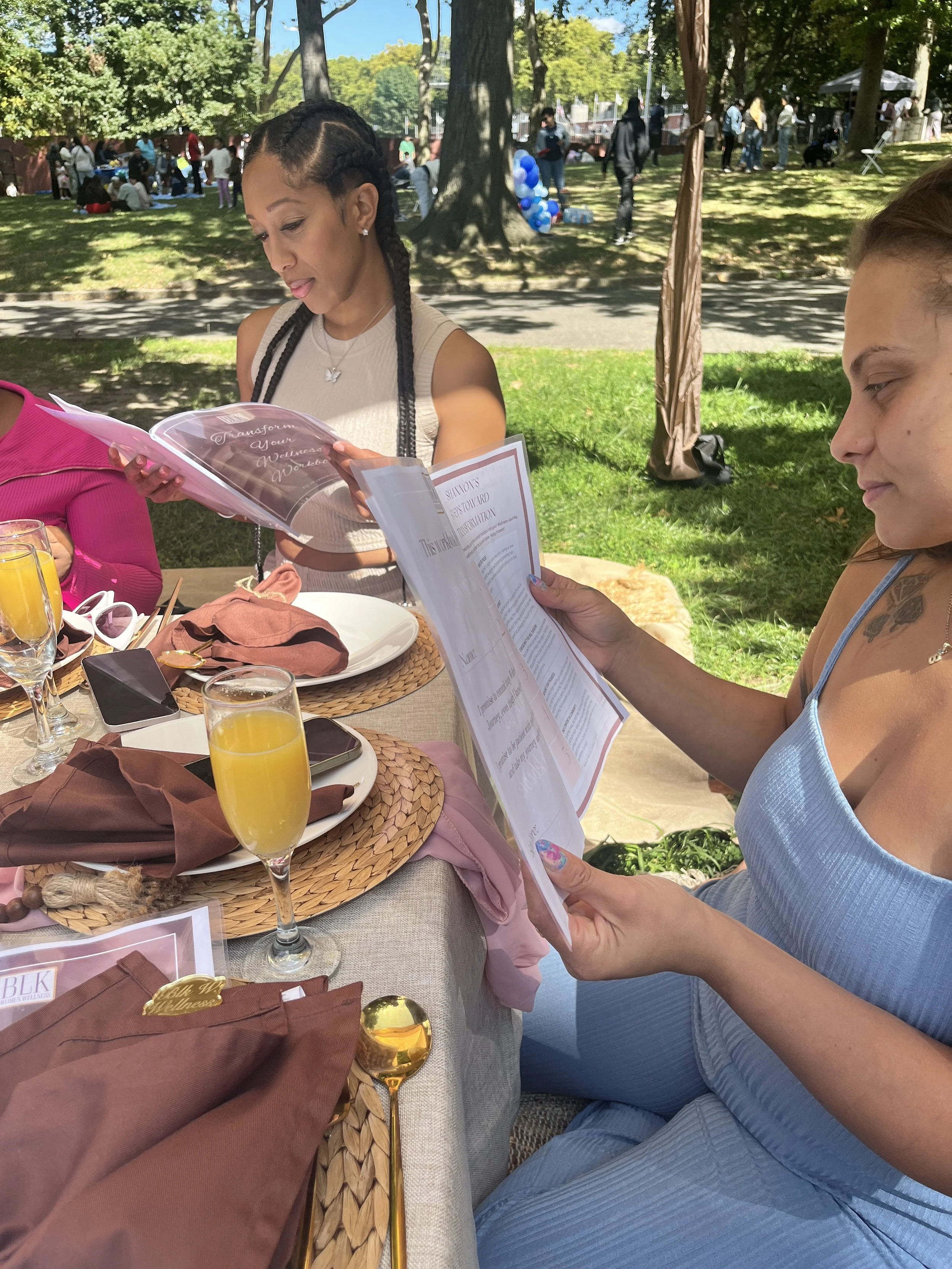 Two women sitting at an outdoor table with menus, drinks, and place settings, looking at their menus during a gathering in a park.