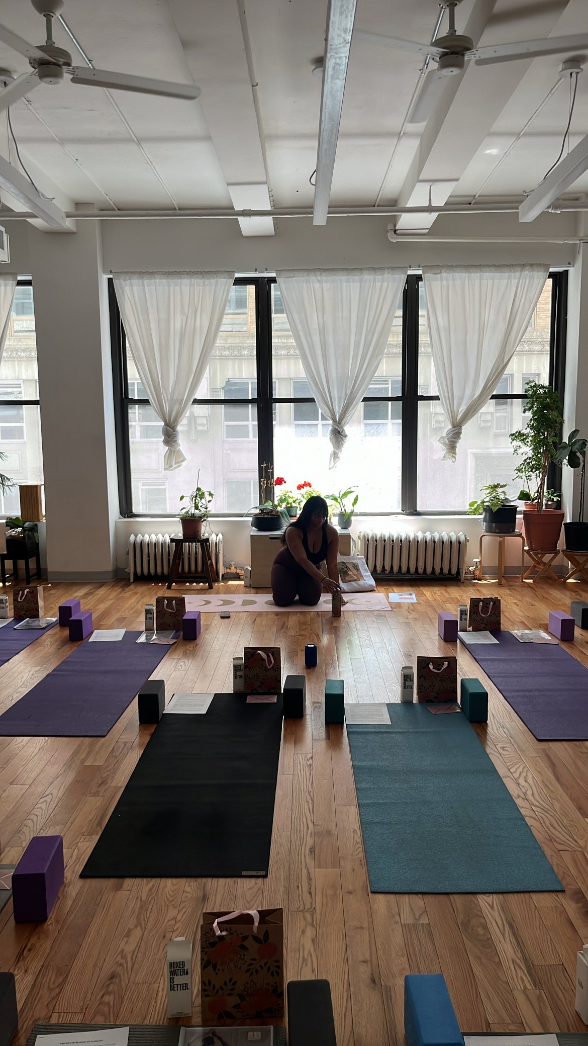 A woman practicing yoga in a well-lit studio with wooden floors, large windows with white curtains, and potted plants. The studio is set up with yoga mats, blocks, and gift bags.