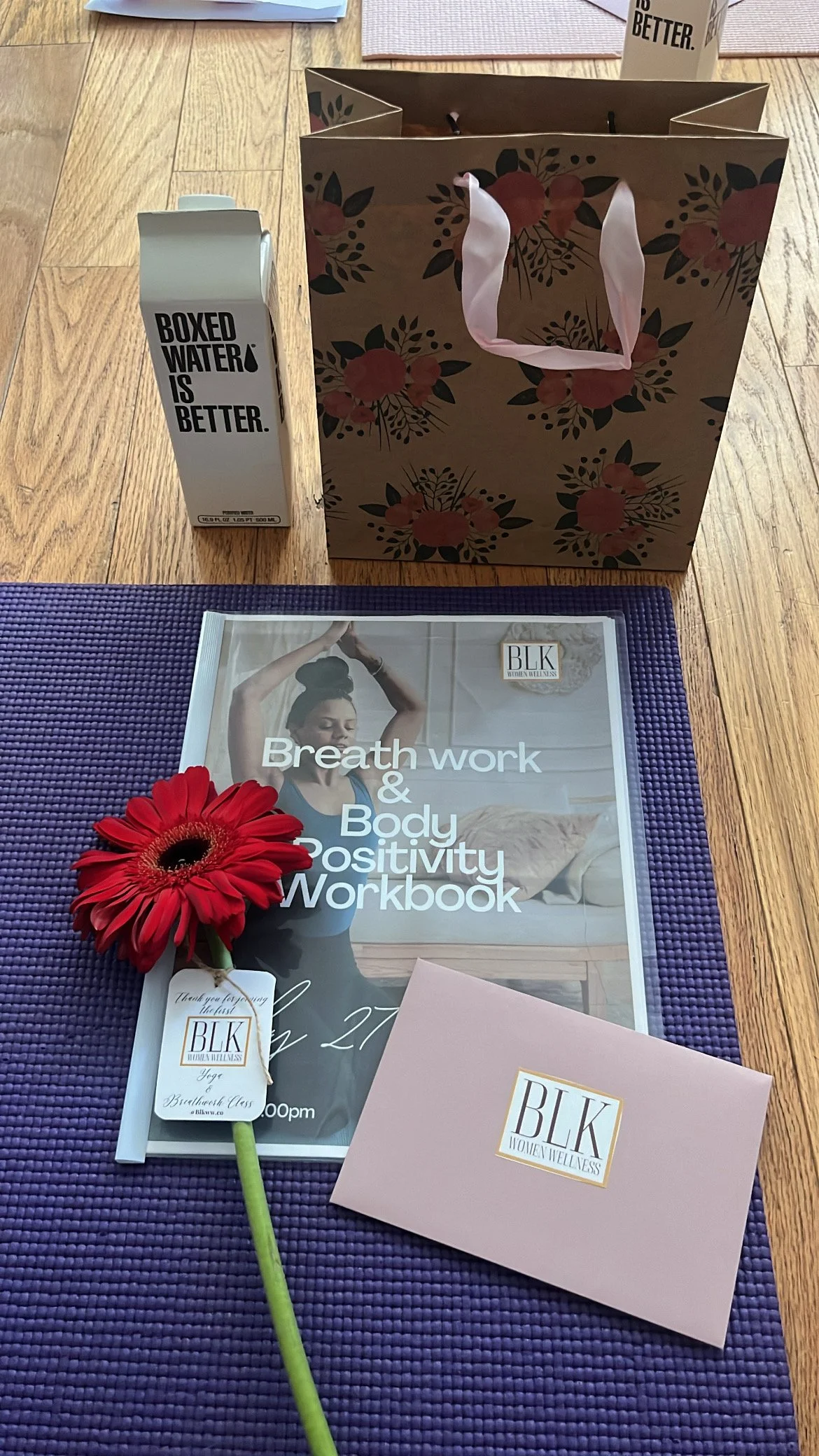 A gift bag with floral design, a box of boxed water, a workbook titled 'Breath work & Body Positivity Workbook,' a pink card with a logo 'BLK Women Wellness,' a red flower with a tag that says 'Thank you for paying the best,' and a purple mat on a wo