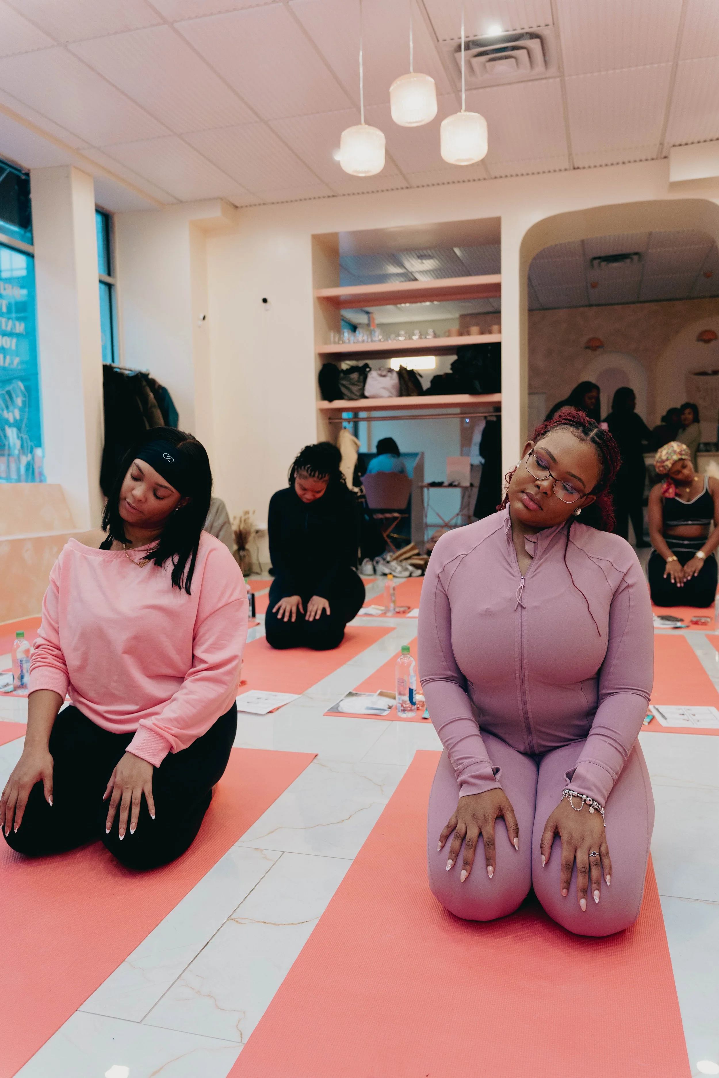 Group of women participating in a yoga or meditation class, seated on pink mats with water bottles, in a well-lit indoor space with white walls and ceiling lights.