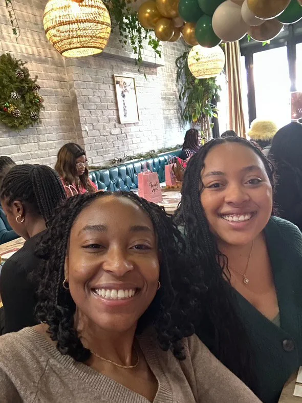 Two young women smiling at a table in a decorated restaurant with balloons, a wreath, and string lights hanging from the ceiling.