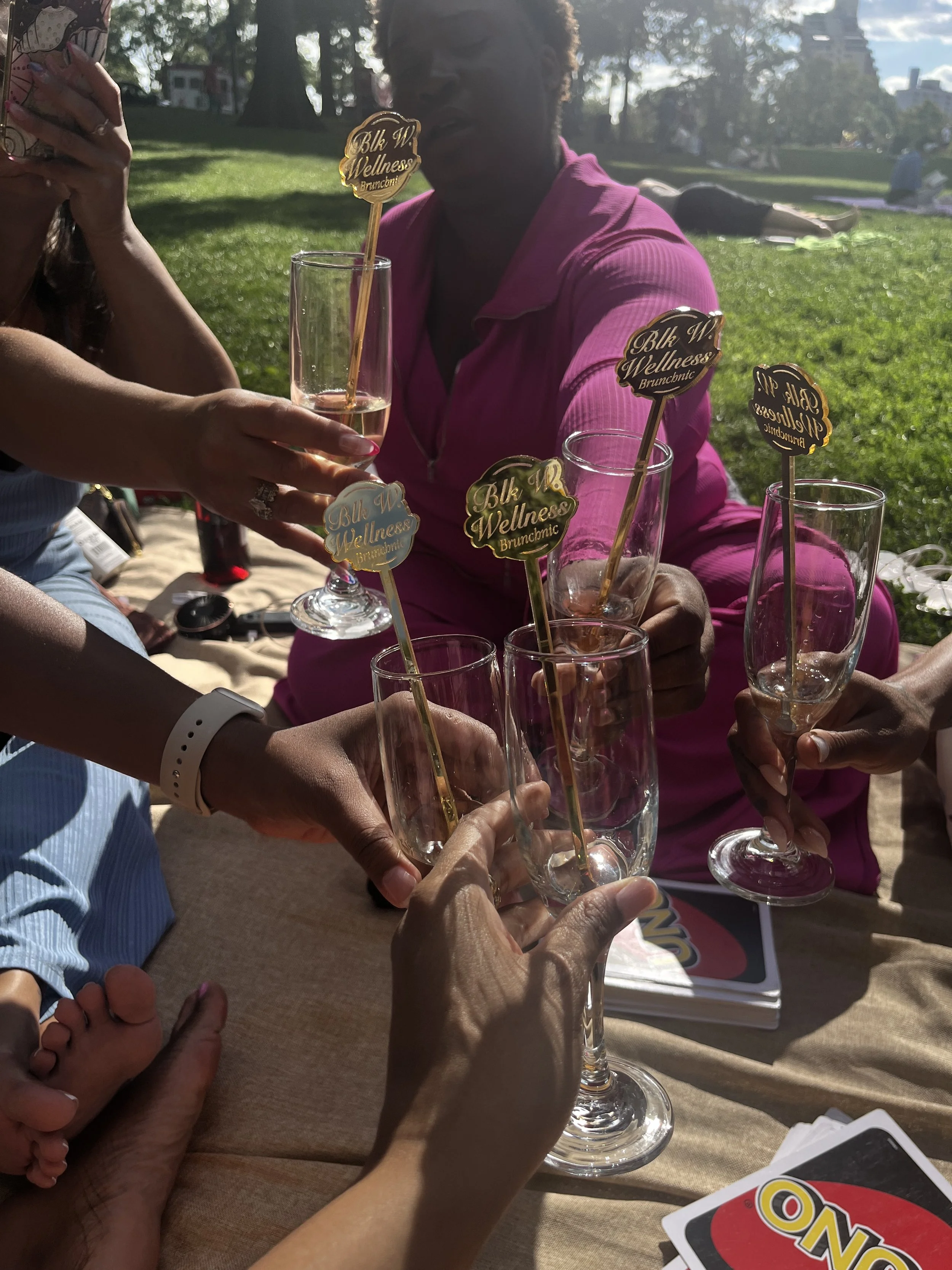 Group of people celebrating outdoors with champagne glasses and event markers that read 'Blk W Wellness Brunch'.