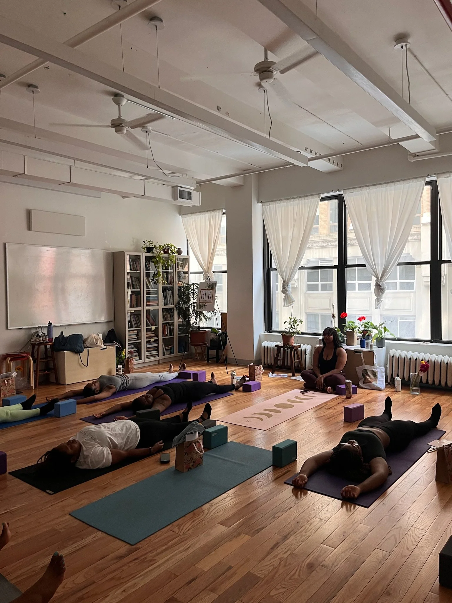 A group of people participating in a yoga class in a bright studio with large windows, wooden floors, and potted plants. They are lying on yoga mats in a relaxed position, with an instructor sitting cross-legged nearby.