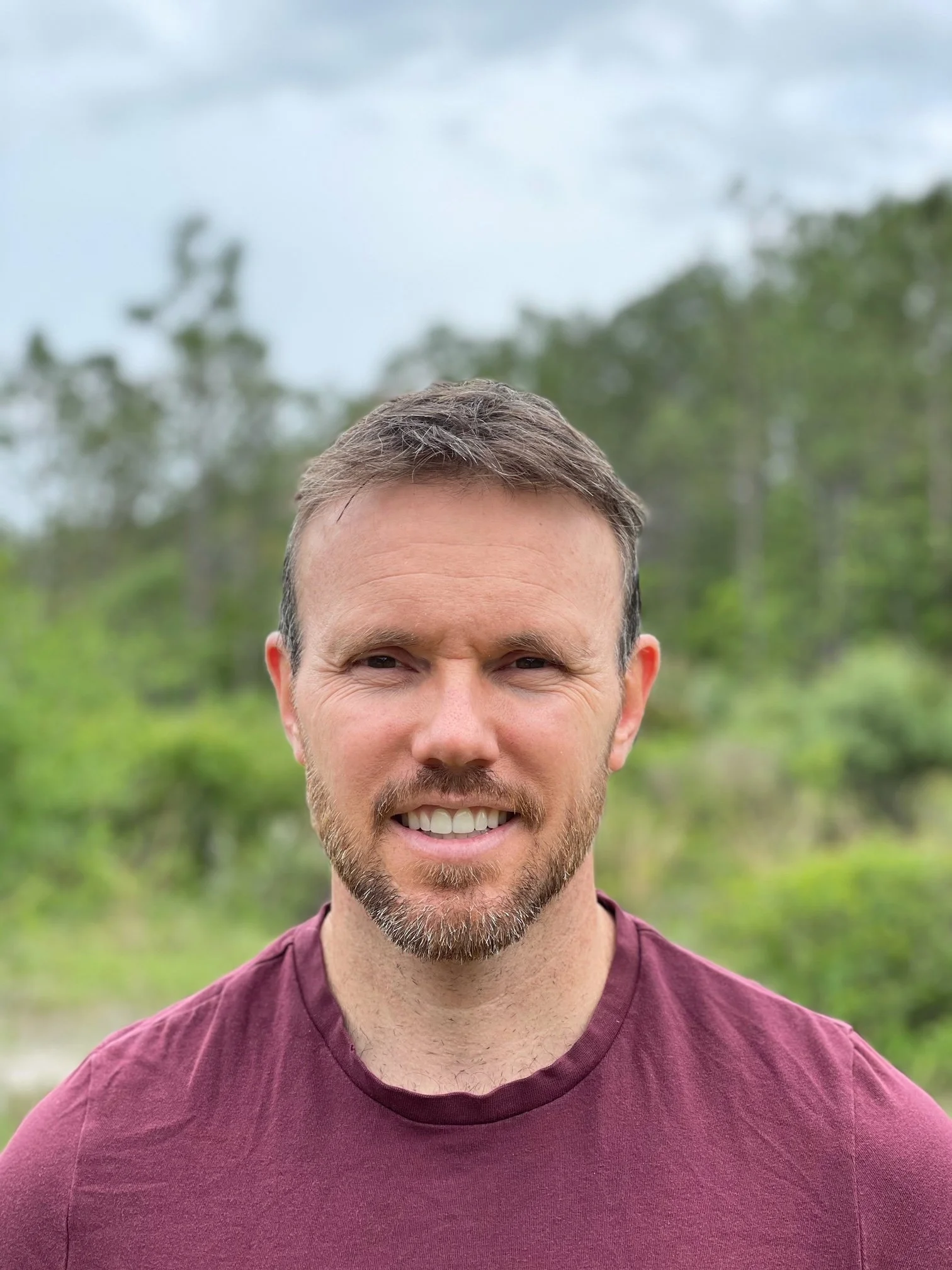 A man with short brown hair and a beard, smiling outdoors with green trees and cloudy sky in the background.