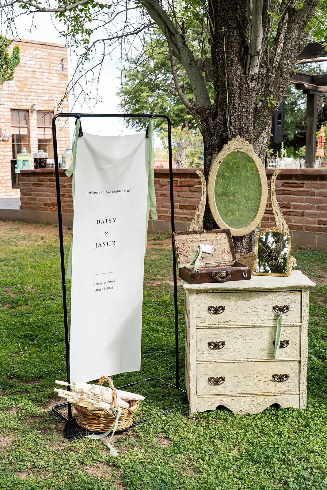 Outdoor wedding setup with a vintage dresser, mirrors, and a welcome sign for Daisy and Jasur on a grassy lawn near a brick wall and tree at Agua Linda Farm in Amado, AZ