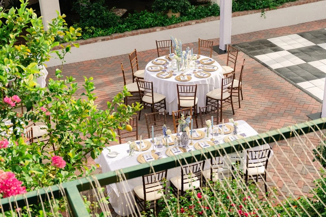 Outdoor patio with two round tables and one rectangular table set for a formal event, decorated with white tablecloths, floral centerpieces, candles, and gold-rimmed plates, surrounded by wooden chairs with beige cushions, and lush greenery and pink flowers nearby