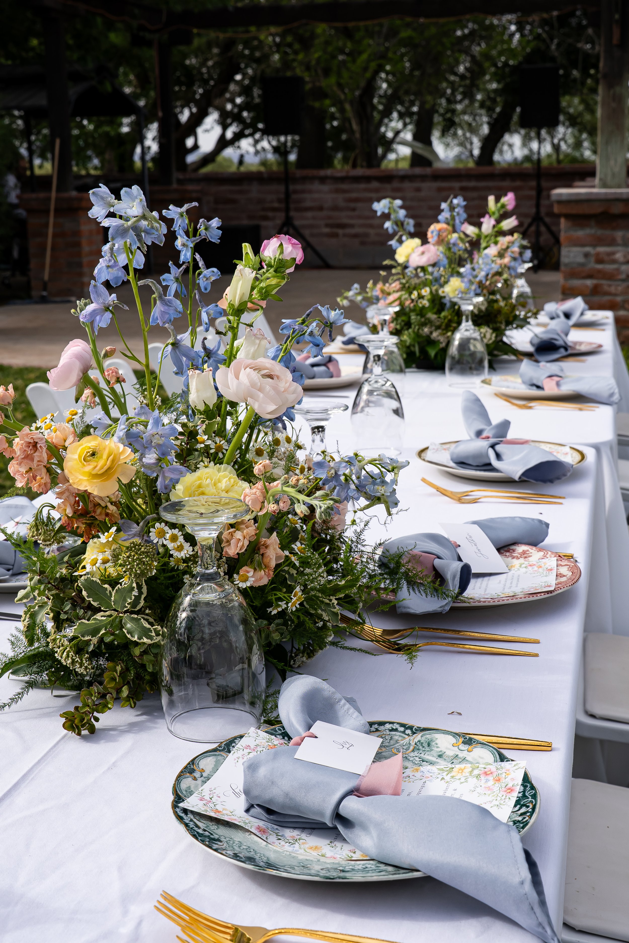 A decorated outdoor dining table with a floral centerpiece, set with plates, napkins, gold utensils, and wine glasses, with greenery and trees in the background at Agua Linda Farm in Amado, Arizona