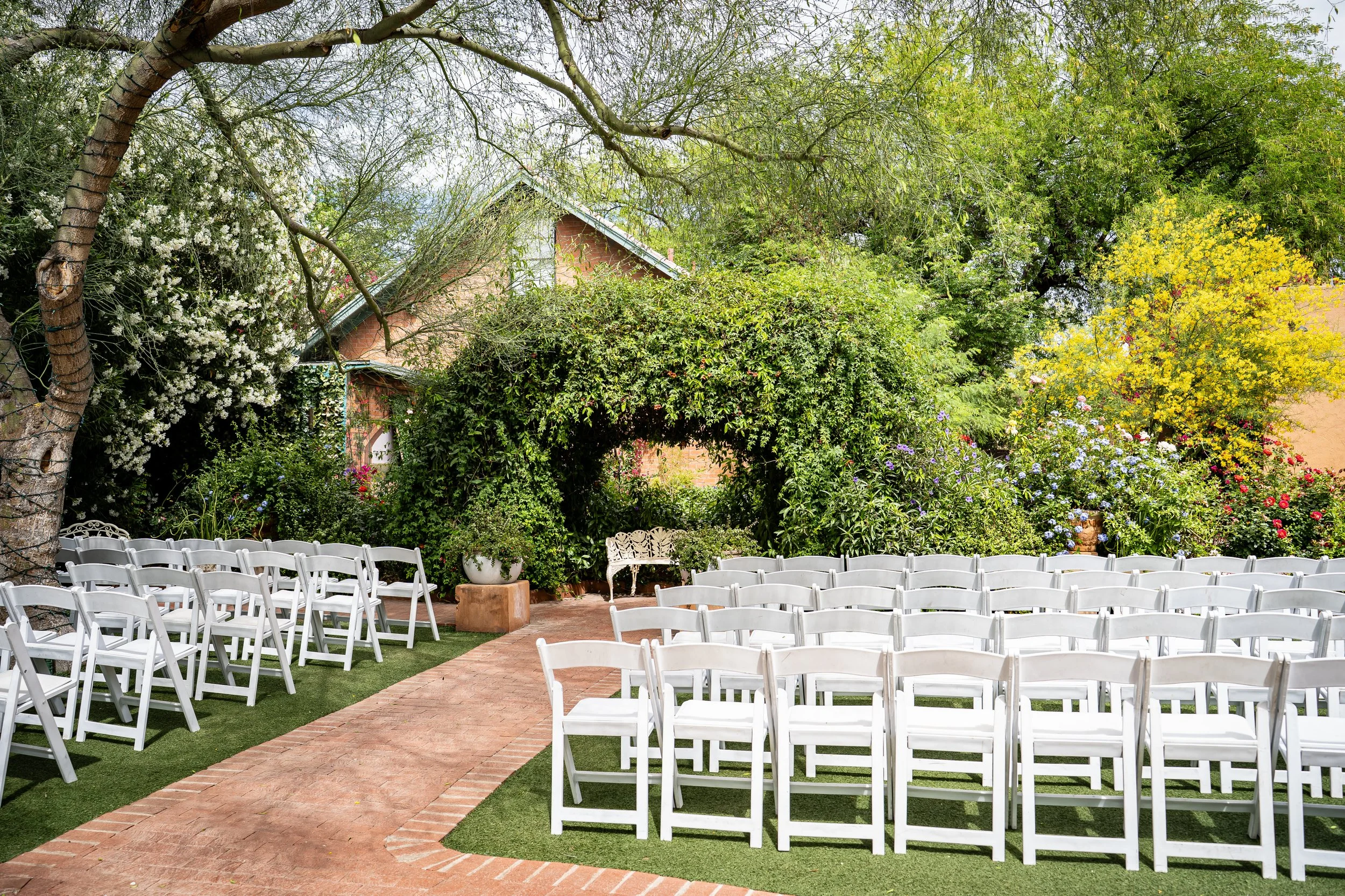 Outdoor wedding or event setup with white folding chairs arranged in rows on a lawn, with a brick walkway, surrounded by lush green trees and blooming flowers at Kingan Gardens in Tucson, Arizona