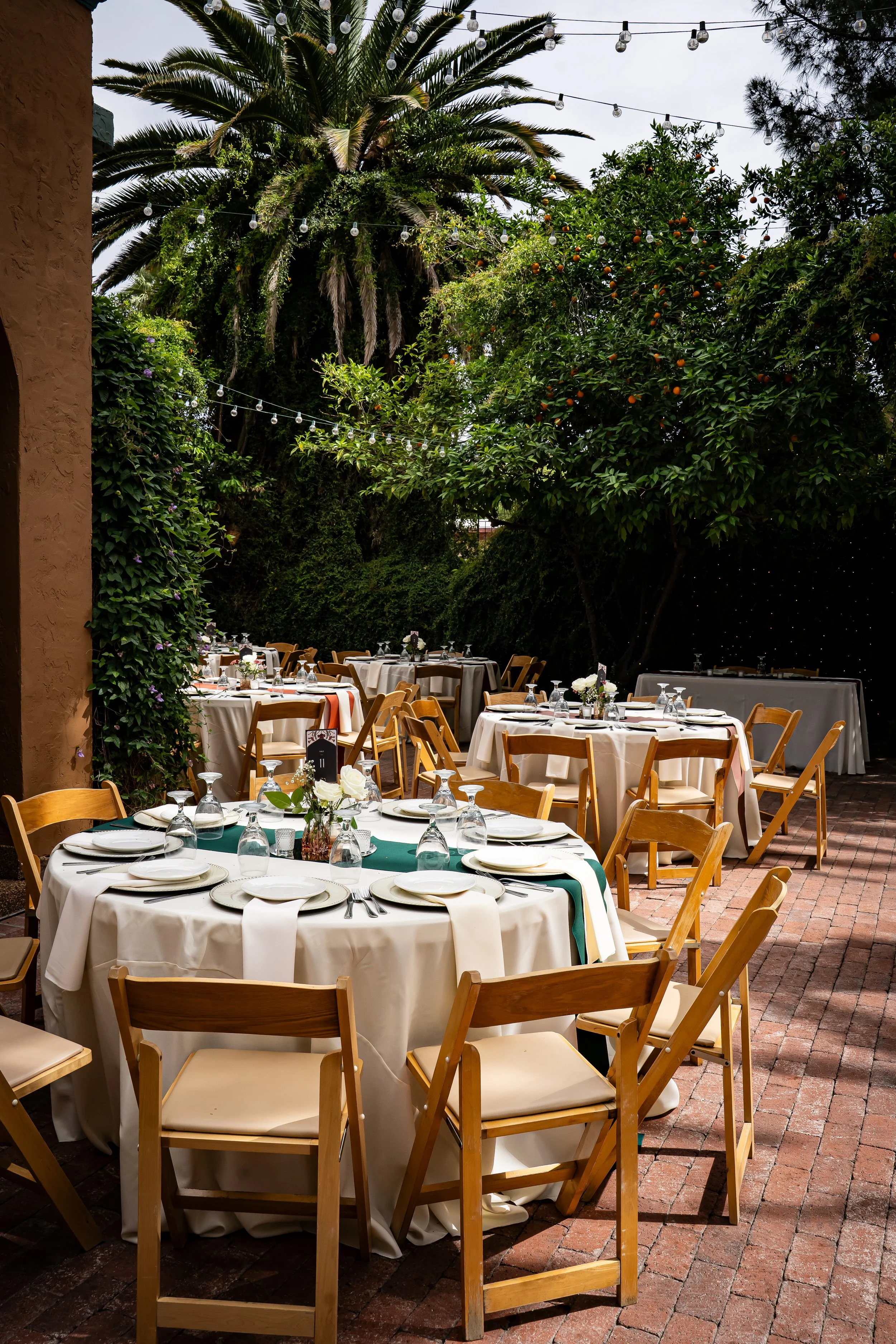 Outdoor restaurant setting with several tables covered in white tablecloths and wooden chairs. Some tables have floral centerpieces and place settings. The area is surrounded by lush green trees and string lights hanging above at Kingan Gardens