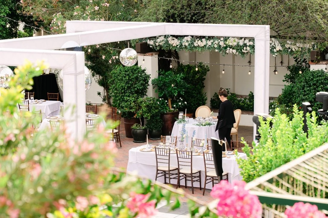 An outdoor event setup with tables decorated with white tablecloths and chairs, under a white canopy frame with hanging lights, surrounded by green plants and pink flowers, with a person arranging the table at Stillwell House in Tucson, Arizona