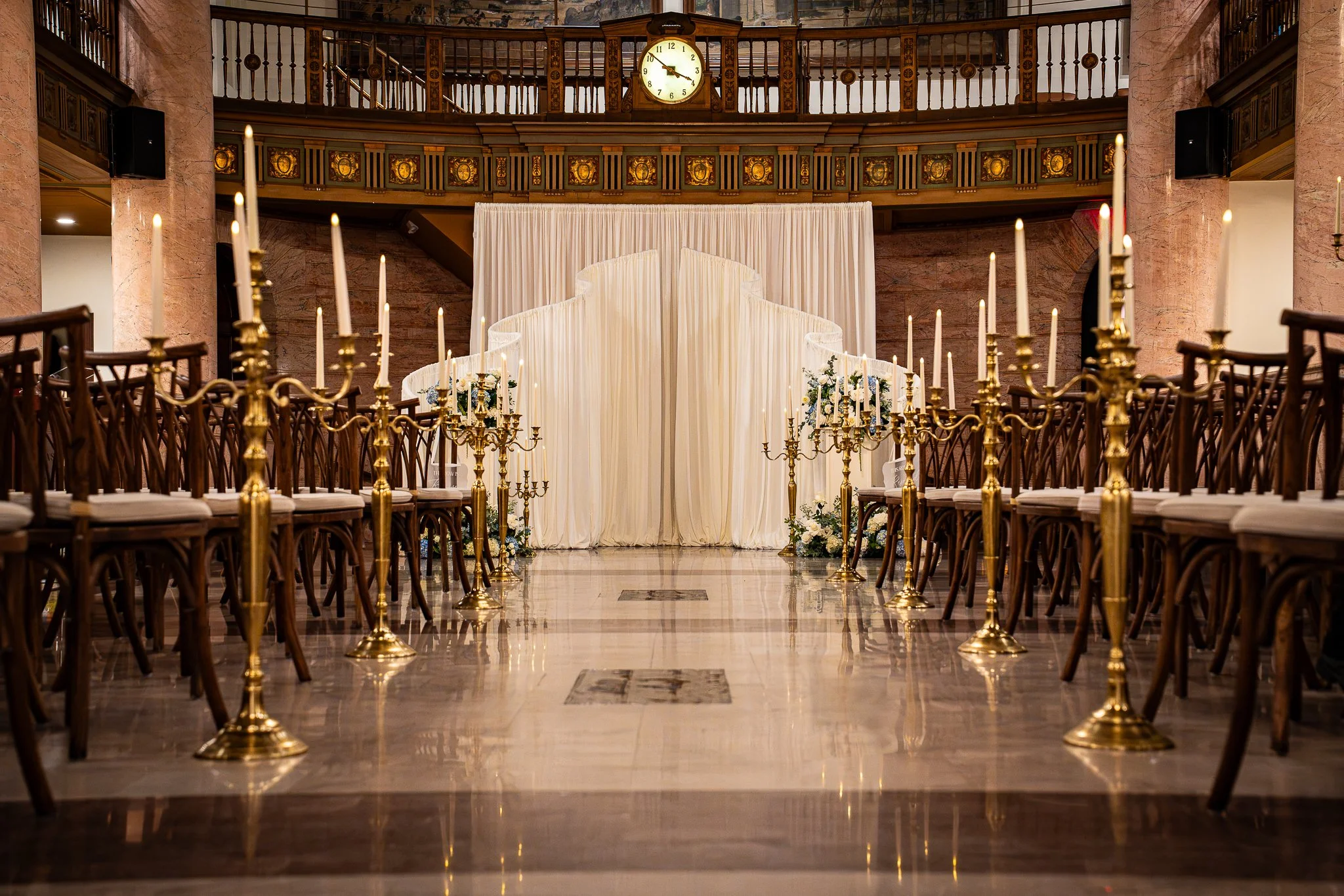 An ornate indoor space set up for a wedding ceremony with rows of chairs, gold candelabras, and a draped aisle with floral arrangements at the end. There is a large clock showing 9:12 and a decorative railing with a mural in the upper background