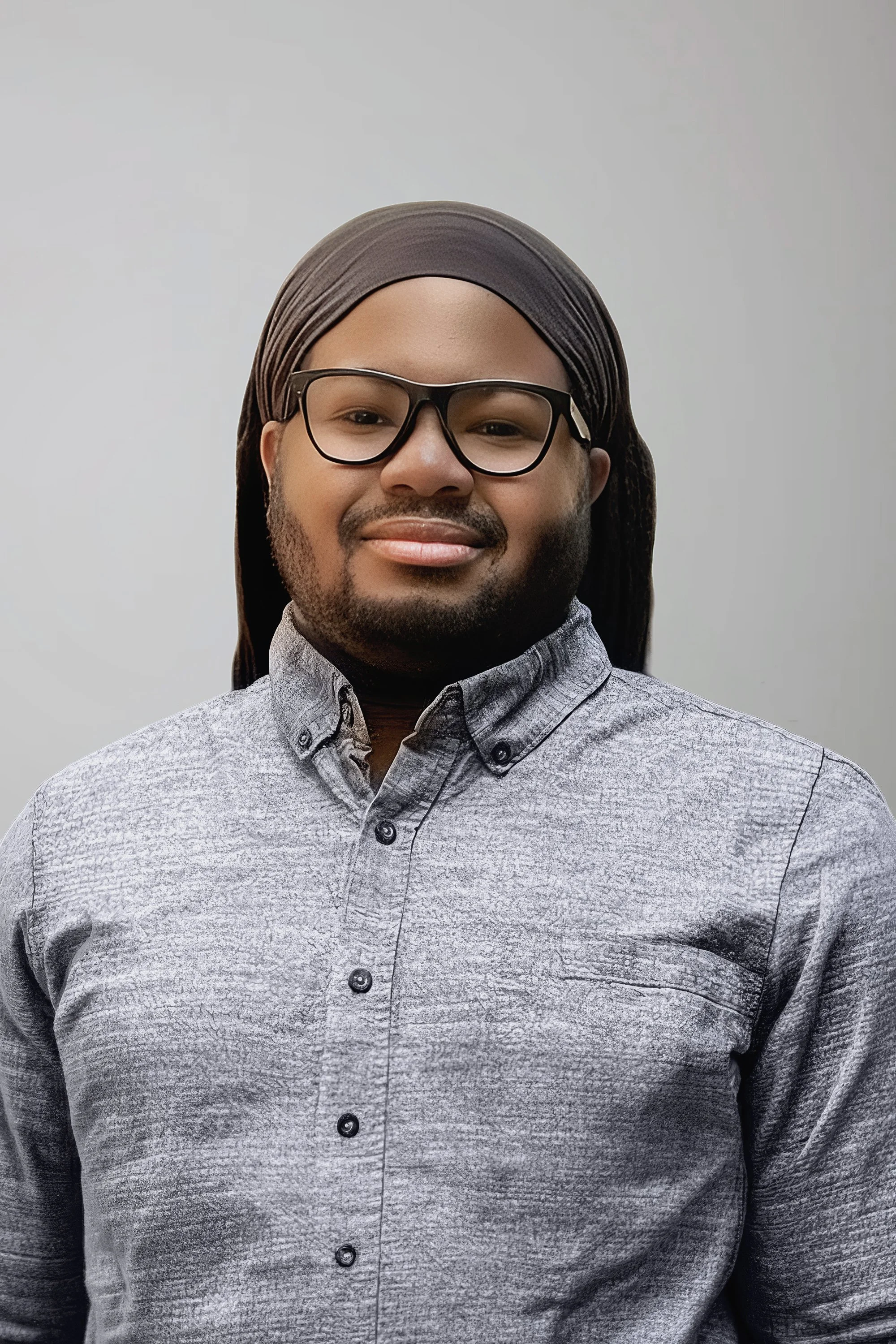 A man with glasses, a beard, and mustache, smiling, wearing a gray patterned button-up shirt and a brown durag, standing against a plain gray background.
