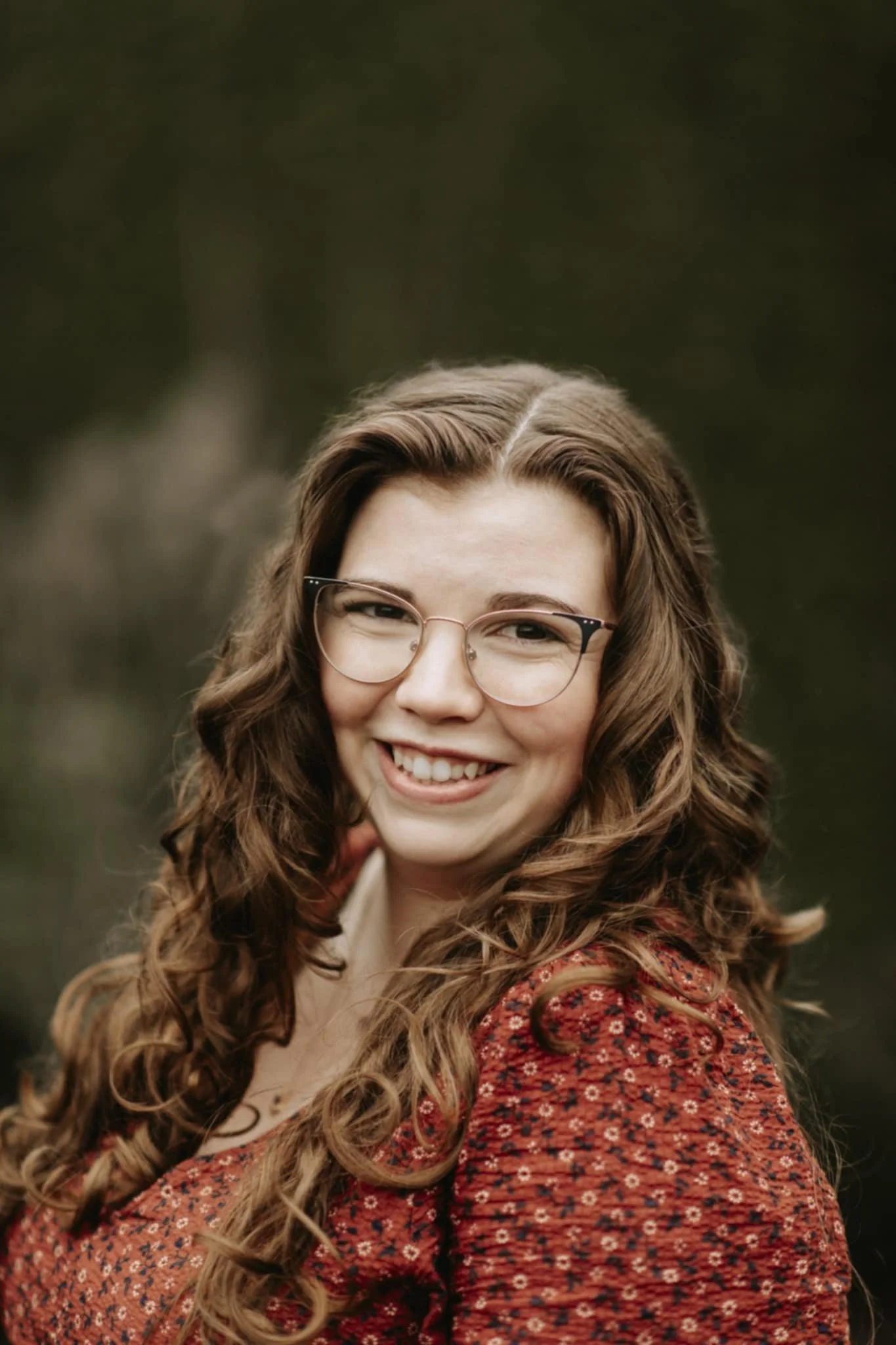 A young woman with curly hair wearing glasses and a red patterned top, smiling outdoors.