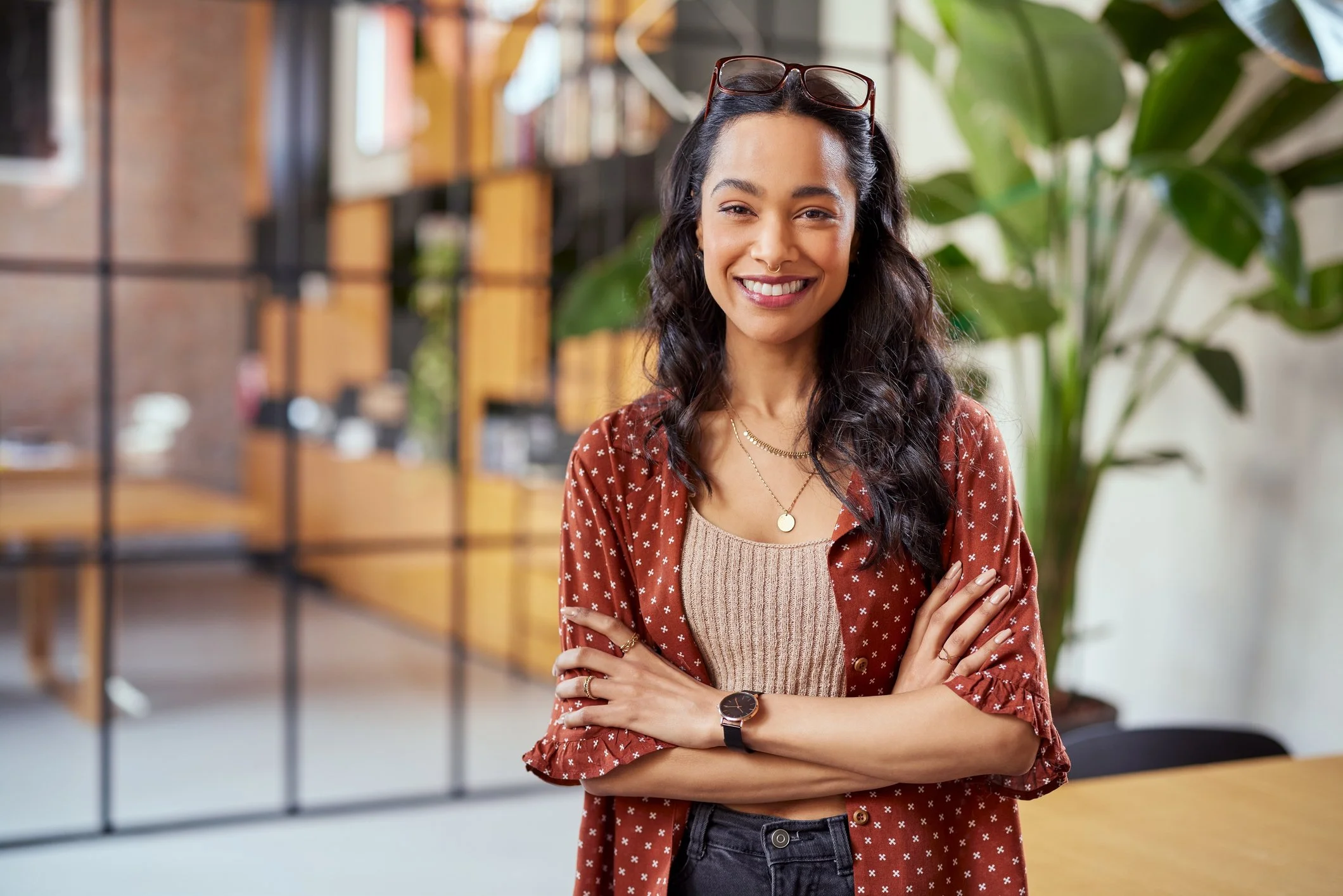 A smiling young woman standing with crossed arms in a modern office space, wearing glasses on her head, layered necklaces, a watch, a beige top, and a red patterned shirt, with green plants and office decor in the background.