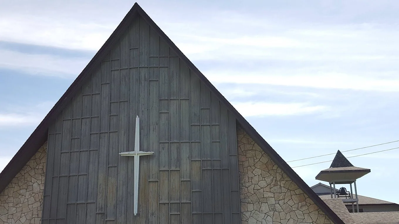 The front of a church with a large cross on its wooden facade, stone accents on the walls, and a small bell tower with a pointed roof.