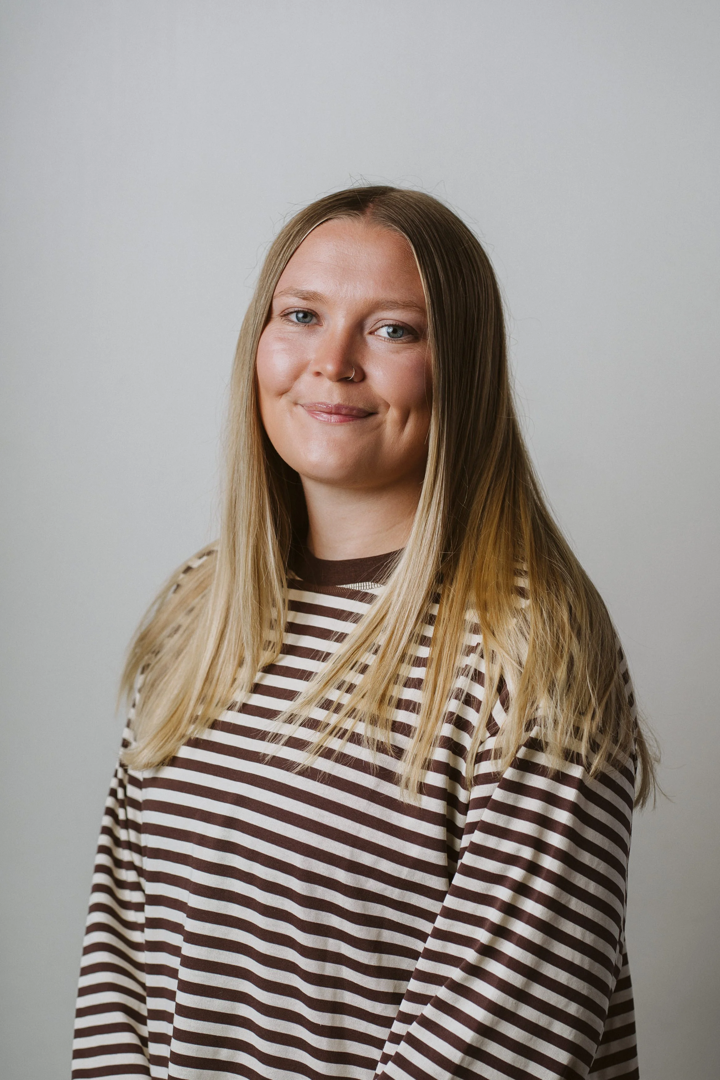 A young woman with long blond hair, blue eyes, and a nose piercing, wearing a brown and white striped shirt, stands against a plain gray background.